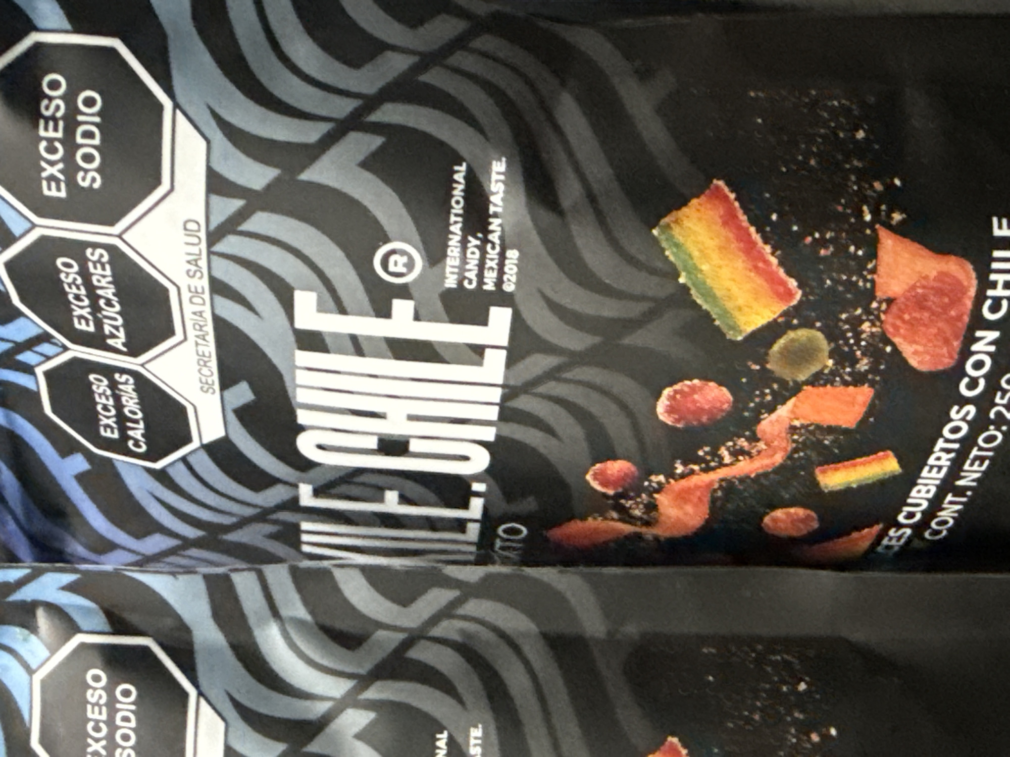 The image shows two packages of "Dato Ile Chile" brand candy. The packages are black with grey wavy patterns. The candy is described as "International Candy, Mexican Taste" and "Ces Cubiertos con Chile" (covered with chili). Each package includes the warning labels "Exceso Sodio" (Excess Sodium), "Exceso Azúcares" (Excess Sugars), and "Exceso Calorías" (Excess Calories), required by Secretaria de Salud (Secretary of Health). There is no geographical information available about this image so I am unable to provide a location in the city of San Bernardino Tlaxcalancingo, Mexico.