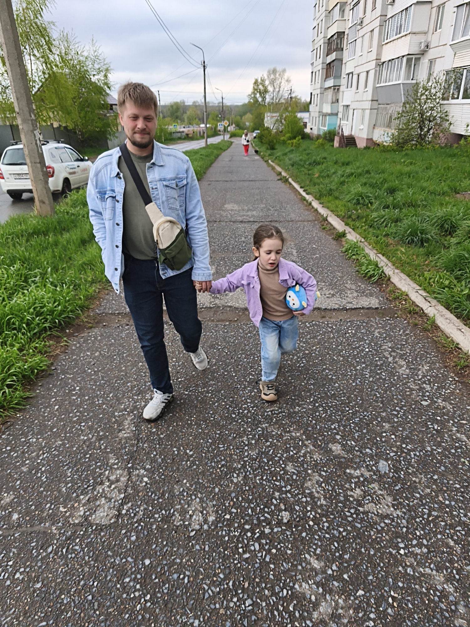 A man and a young girl are walking hand-in-hand on a paved sidewalk. The man is wearing a light blue denim jacket, a dark green shirt, dark jeans, and white sneakers. He has a light-colored cross-body bag. The girl is wearing a light purple jacket, a brown shirt, light blue jeans, and brown shoes. She is holding a blue plush toy in her other hand. They are walking away from the camera.
In the background, there is a residential building on the right side of the sidewalk and a road with a white car on the left. In the distance, there is another person walking on the sidewalk. The sky is overcast.
Based on visual features, including building styles and vehicle license plates, the location is likely in the city of Chaikovsky, Russia.