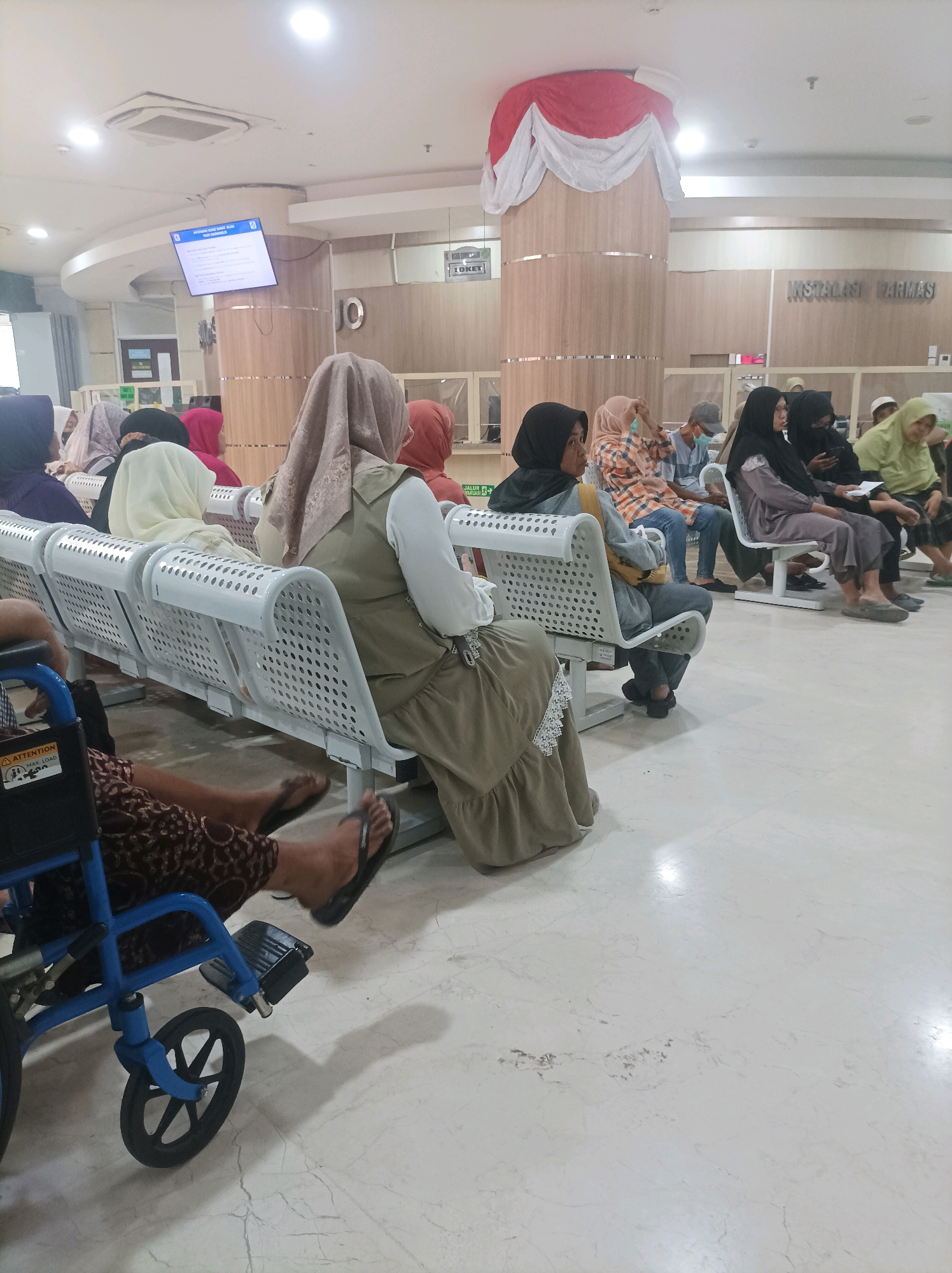In a waiting area, likely within a medical facility in Sumuragung, Indonesia, several people are seated on white benches. A woman in a wheelchair, wearing a patterned brown skirt and black flip-flops, is positioned in the foreground to the left. The benches are arranged in rows and appear to be made of white perforated metal. Above the waiting area, a large screen displays information, and a sign indicates "INSTALASI FARMASI." Decorative red and white fabric hangs from a pillar in the background. The floor is tiled with a marble-like pattern.