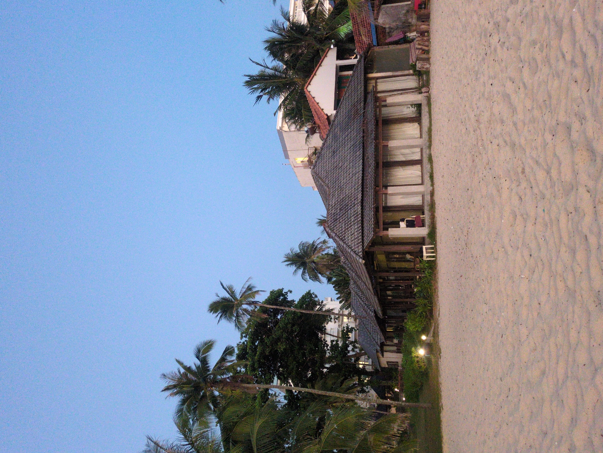 The image captures a beachfront scene in Phú Quốc, Vietnam. A long, low-lying building with a dark, tiled roof stands parallel to the sandy beach. The building's facade features numerous windows. A row of lush greenery separates the building from the beach. Palm trees are visible against a light blue sky. In the background, a taller building can be seen behind the lower structure. The sand is light in color and appears relatively undisturbed.