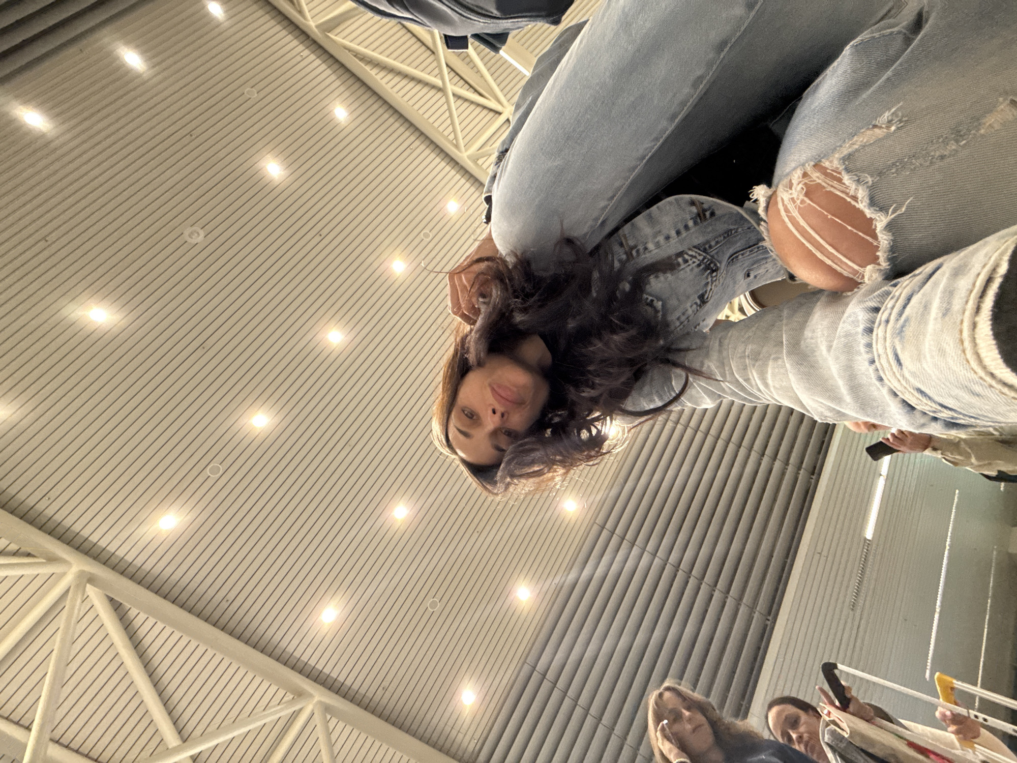 The image, taken from a low angle, shows a woman with long, wavy brown hair, wearing a denim jacket and ripped jeans, posing for the camera. In the background, partially visible, are other people with luggage, suggesting an airport setting. The ceiling is a distinctive metal structure with linear patterns and inset lighting. Given these elements, the location is likely Sofia Airport, Bulgaria.