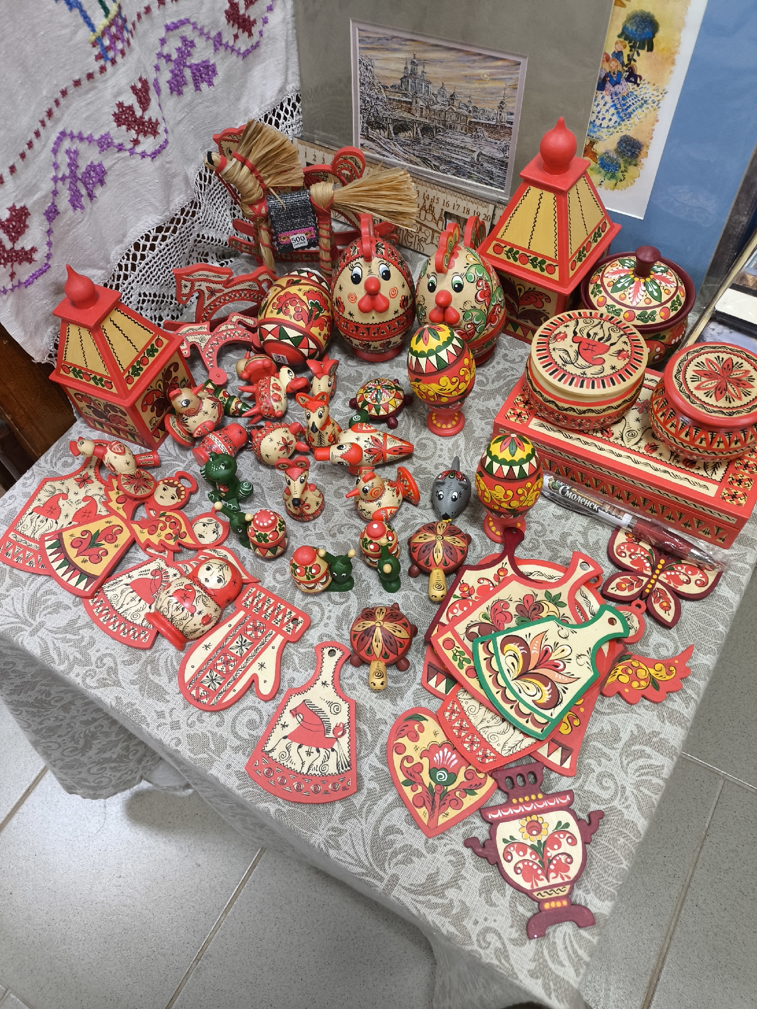 A high-angle shot captures a collection of intricately painted wooden crafts arranged on a table. The crafts are predominantly red, yellow, green, and white, and feature traditional Russian folk art designs. Several items are shaped like eggs, while others resemble animals, buildings, and kitchen utensils. Some pieces are stacked, creating varying heights across the tableau.
In the background, a white cloth with colorful embroidery adds a textural element to the scene. Framed artwork stands behind the crafts, providing a glimpse into the environment, although the specific city or location remains unidentified. The table is covered with a patterned tablecloth, and the floor is tiled with neutral-colored squares. A pen marked "СМОЛЕНСК" (Smolensk) is visible, suggesting the crafts may be from or related to the city of Smolensk.