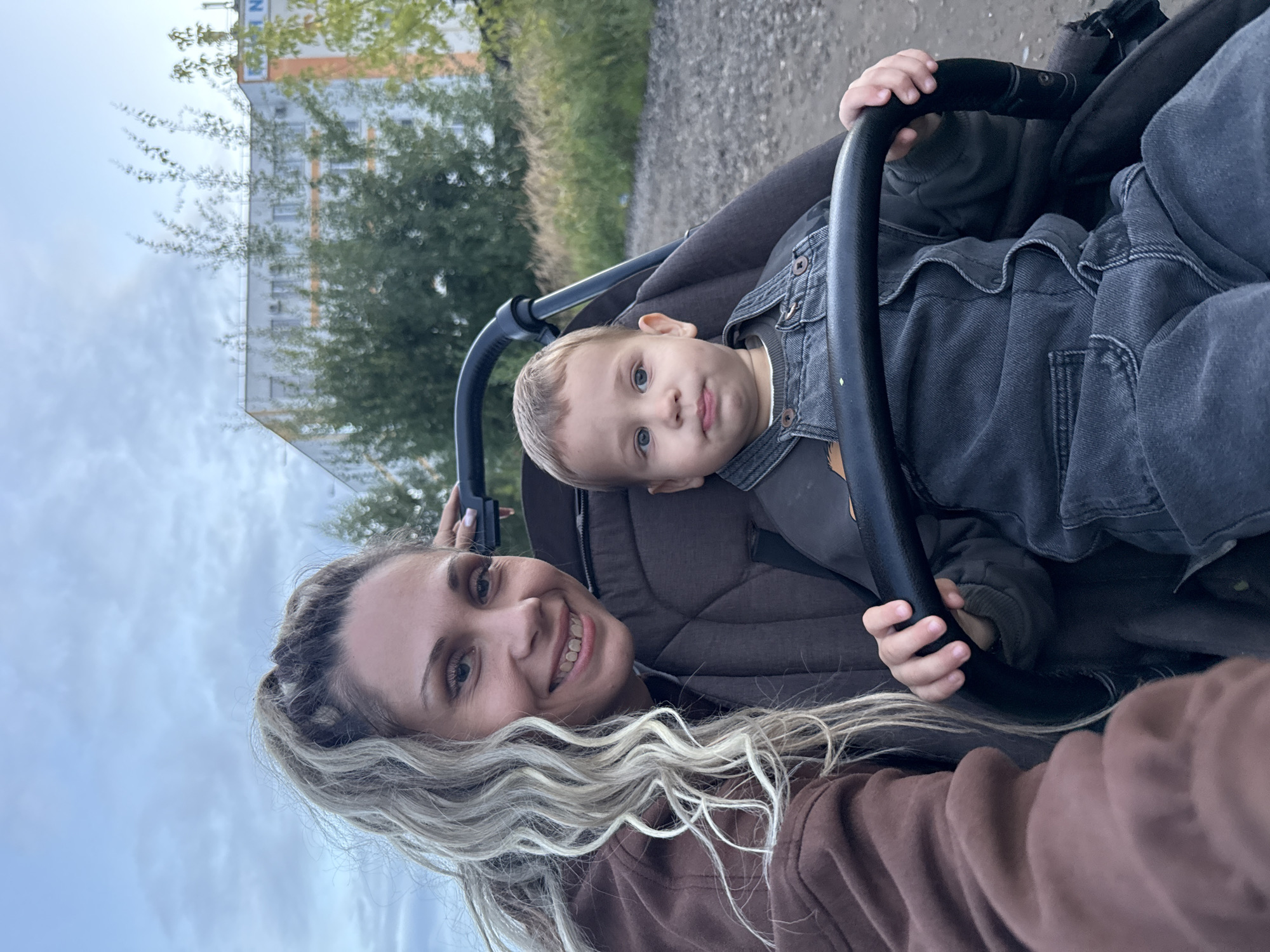 A woman and a young boy are seated in a stroller. The woman, with blonde braided hair and a brown hoodie, is smiling at the camera. The boy, wearing denim overalls, is looking forward with a slightly pensive expression. The stroller is positioned on a gravel path, with trees and a building visible in the background. The setting appears to be an outdoor area, possibly a park or residential street, in Rusanovo, Russia, given the presence of the building and vegetation typical of the region. The sky is overcast, suggesting it might be late afternoon or early evening.