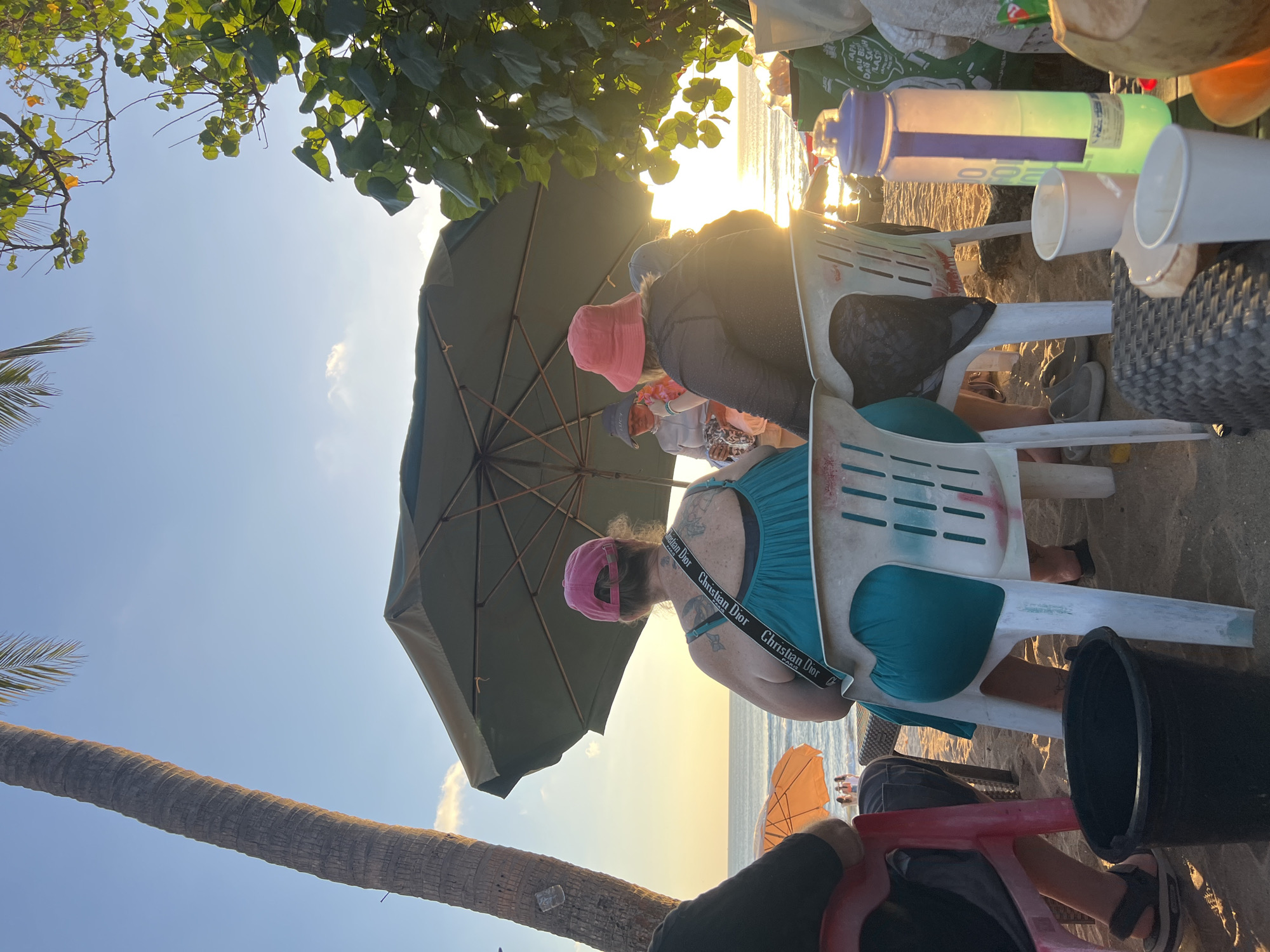 This image depicts a beach scene at sunset in Kuta, Indonesia. A group of people are seated on plastic chairs under a large umbrella on the sandy beach. The sky is clear with a few clouds, and the sun is low on the horizon, casting a warm glow. In the foreground, a black bucket sits on the sand. To the right, a water bottle and several cups are visible on a woven surface, alongside what appears to be a coconut. In the background, the ocean stretches out to meet the sky, with a few other beach umbrellas and people visible further down the shore. A tall palm tree stands to the left, and lush green foliage frames the upper right corner of the image.