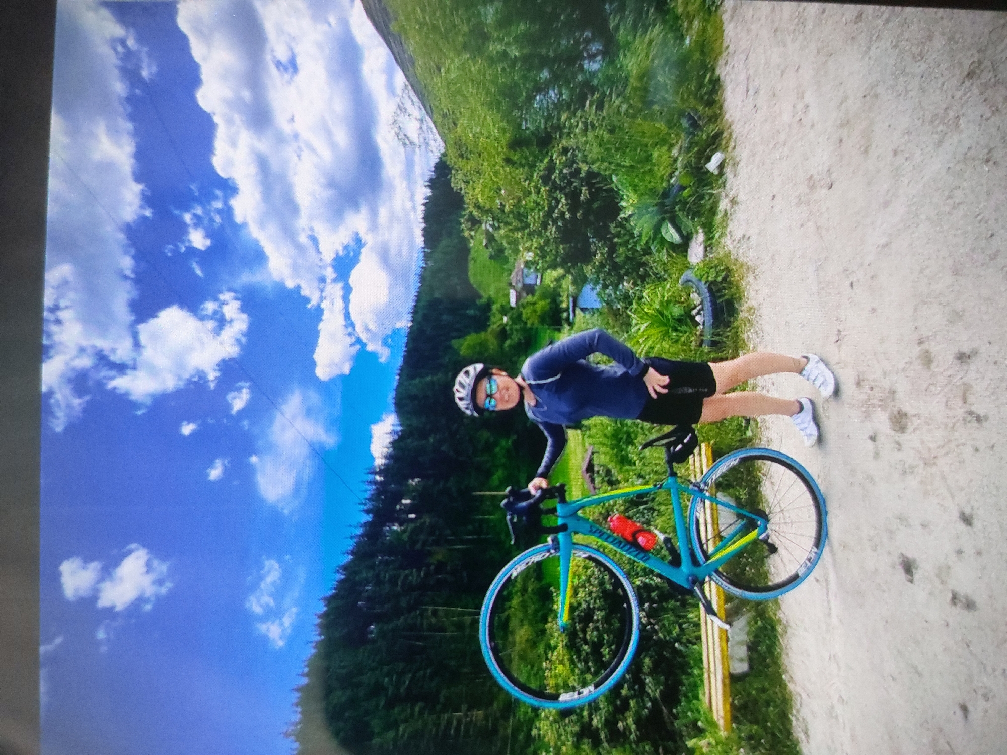 A photograph depicts a cyclist standing beside a turquoise and yellow Specialized bicycle on a path. The cyclist wears a blue long-sleeved shirt, black shorts, a white helmet, and white sneakers. The setting appears to be a mountainous region near Tsarevo, Bulgaria, as the background includes forested hills and a blue sky with clouds. The path is made of light-colored gravel. There is a glimpse of a house between trees.