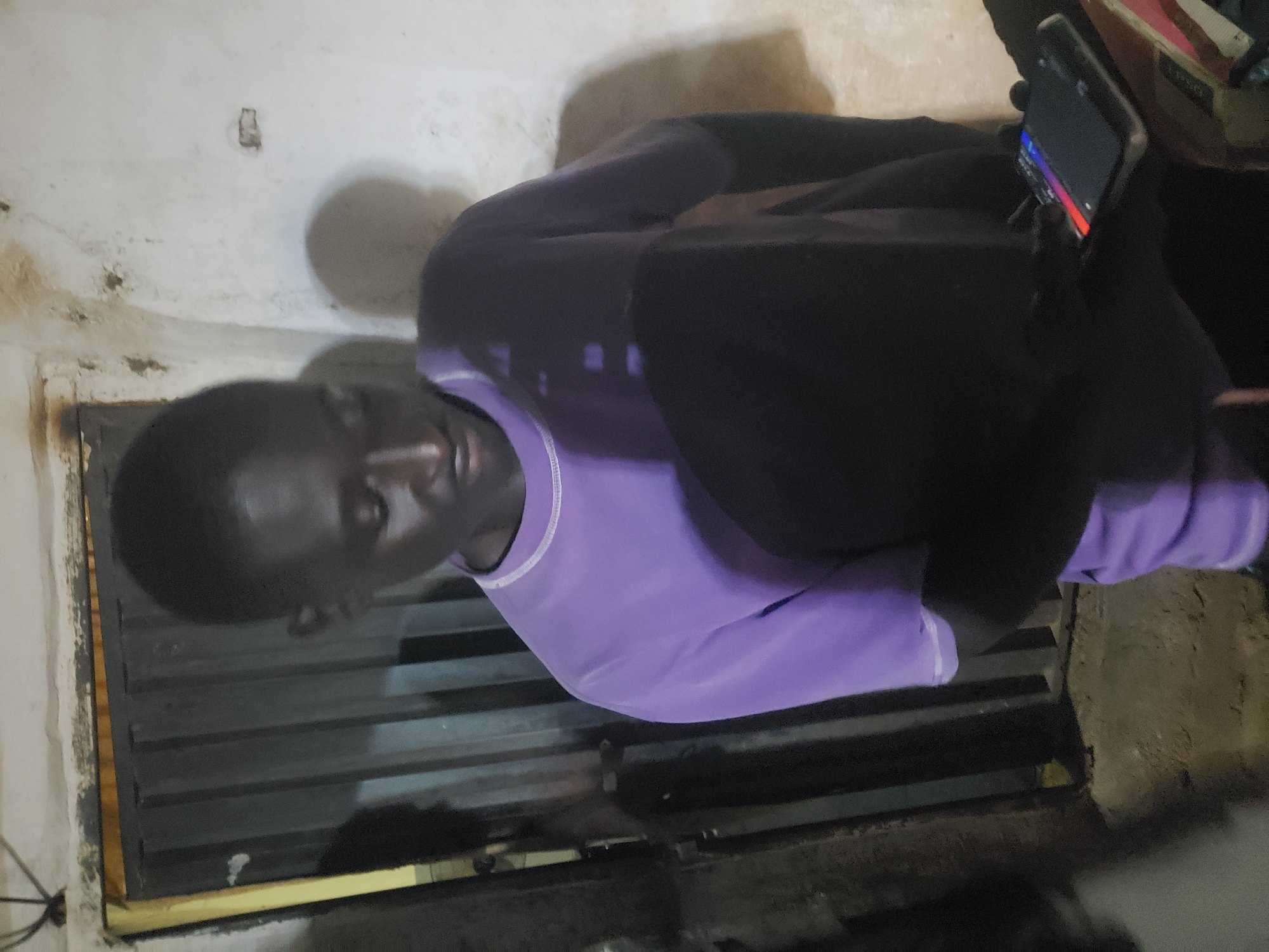 A person is sitting indoors and holding a smartphone. The individual is wearing a lilac-colored shirt with white trim and a dark-colored jacket. The background features a louvered window and a wall. No information within this image can confirm the location as Bukuru, Nigeria.