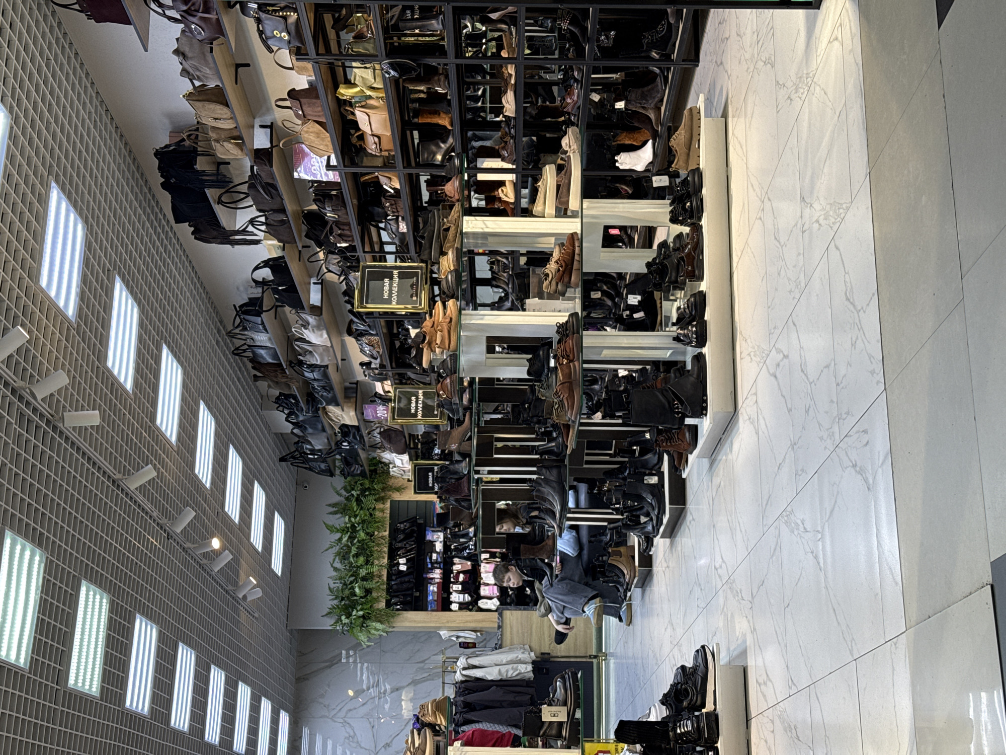 The image displays the interior of a retail store specializing in footwear and handbags, located in Unknown, Unknown. Overhead, a suspended grid ceiling is fitted with numerous rectangular LED light panels and several track spotlights providing bright illumination. The floor is composed of white square tiles with a subtle gray marble-like veining.
The store is dominated by extensive displays of shoes and boots on the lower levels, primarily men's and women's styles in shades of black, brown, and tan. These are arranged on multi-tiered black metal shelving units and white display pedestals. Above the footwear, wall-mounted shelves showcase a wide selection of handbags and shoulder bags, predominantly in dark and neutral colors. Several signs in Russian, including "НОВАЯ КОЛЛЕКЦИЯ" (New Collection) and one indicating a "–20%" discount, are affixed to the displays. Artificial green foliage decorates a recessed area of the back wall. On the far left, a clothing rack holds various garments.
Two individuals are present, seated on a dark bench in the midground, partially obscured by shoe displays. A young male, wearing a dark jacket, light blue jeans, and light-colored sneakers, is seated on the left, looking towards his left. Behind him, a female is also seated, largely obscured from view. Neither individual is actively interacting with the merchandise or with each other.