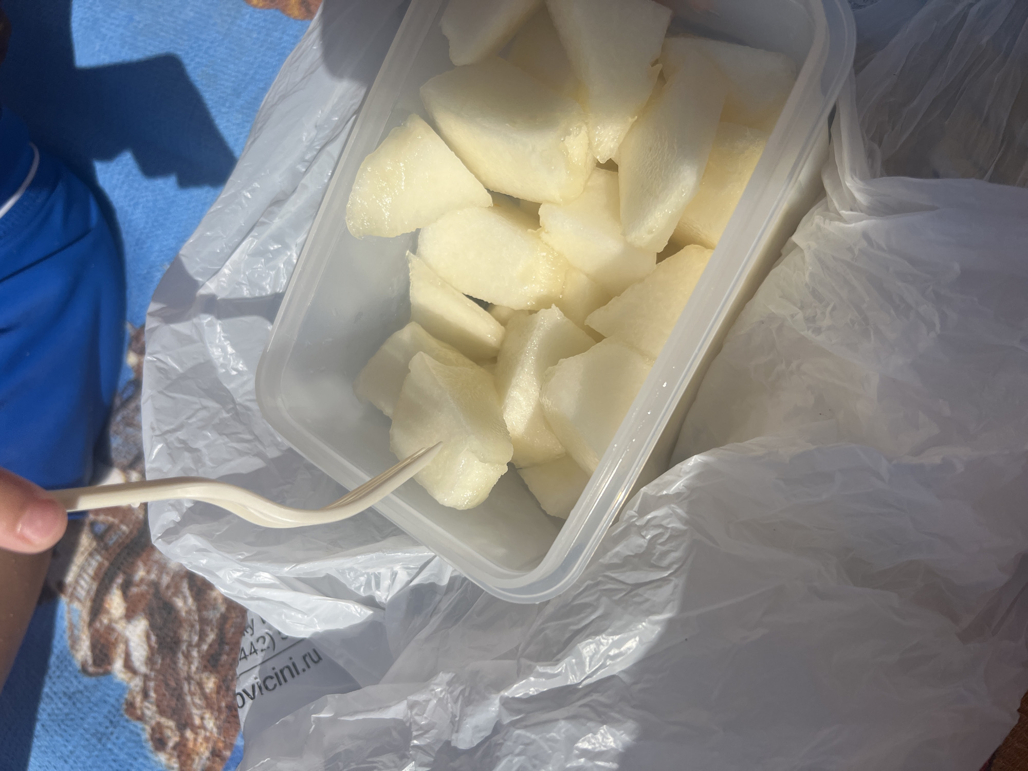 A close-up, overhead shot captures a plastic container filled with diced honeydew melon, resting inside a crumpled white plastic bag. A pale plastic fork is partially submerged in the melon, with a person's hand visible at the top of the frame holding the fork. The background features a blue surface, possibly a beach towel, with a brown and tan patterned object visible on the left. The overall scene suggests an outdoor setting, possibly a picnic or beach outing. The location provided, Kilyakovka, Russia, is not discernible from the visual information.