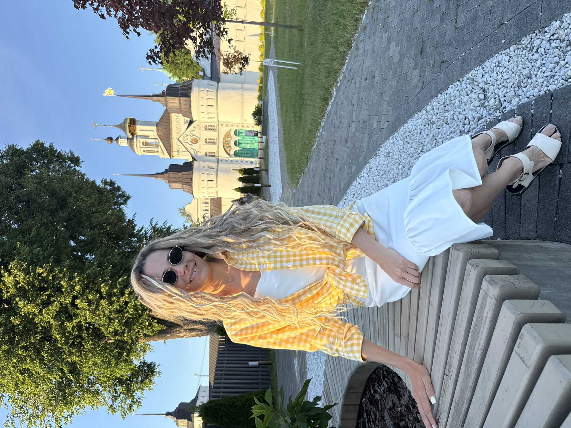 The image shows a woman seated on a curved wooden bench outdoors, with the Tolga Monastery in Yaroslavl, Russia, prominently featured in the background under a clear blue sky.
The woman has long, wavy blonde hair, styled with some interwoven braids or dreadlocks, and wears dark, oval-shaped sunglasses. She smiles directly at the viewer, revealing her teeth. Her attire consists of a white slip dress or camisole under a yellow and white gingham (plaid) button-down shirt, tied at the waist, and a white midi-length skirt with a ruffled hem. Her left hand, adorned with white nail polish, rests on the wooden slats of the bench, while her right hand is in her lap. She wears light-colored, possibly beige or off-white, flat sandals with ankle straps and black soles.
The Tolga Monastery, a large white architectural complex, dominates the background. It features multiple dark conical spires and a central structure topped with a golden dome and a cross. A golden banner, possibly an angel figure, flies from one of the dark spires. The monastery's facade includes numerous small arched windows and a grand entrance with two ornate green arched double doors.
The surrounding environment is well-maintained, featuring grey paver stone pathways bordered by strips of white gravel. Sections of green grass are visible between the pathways and structures. A large, mature deciduous tree with bright green foliage stands to the left behind the woman, and smaller trees with dark red leaves are situated to the right, closer to the monastery wall. A dark fence runs along the left side of the pathway. The scene is illuminated by bright, natural sunlight, indicating late afternoon or early evening, which casts warm highlights on the woman's hair and the monastery's facade.
