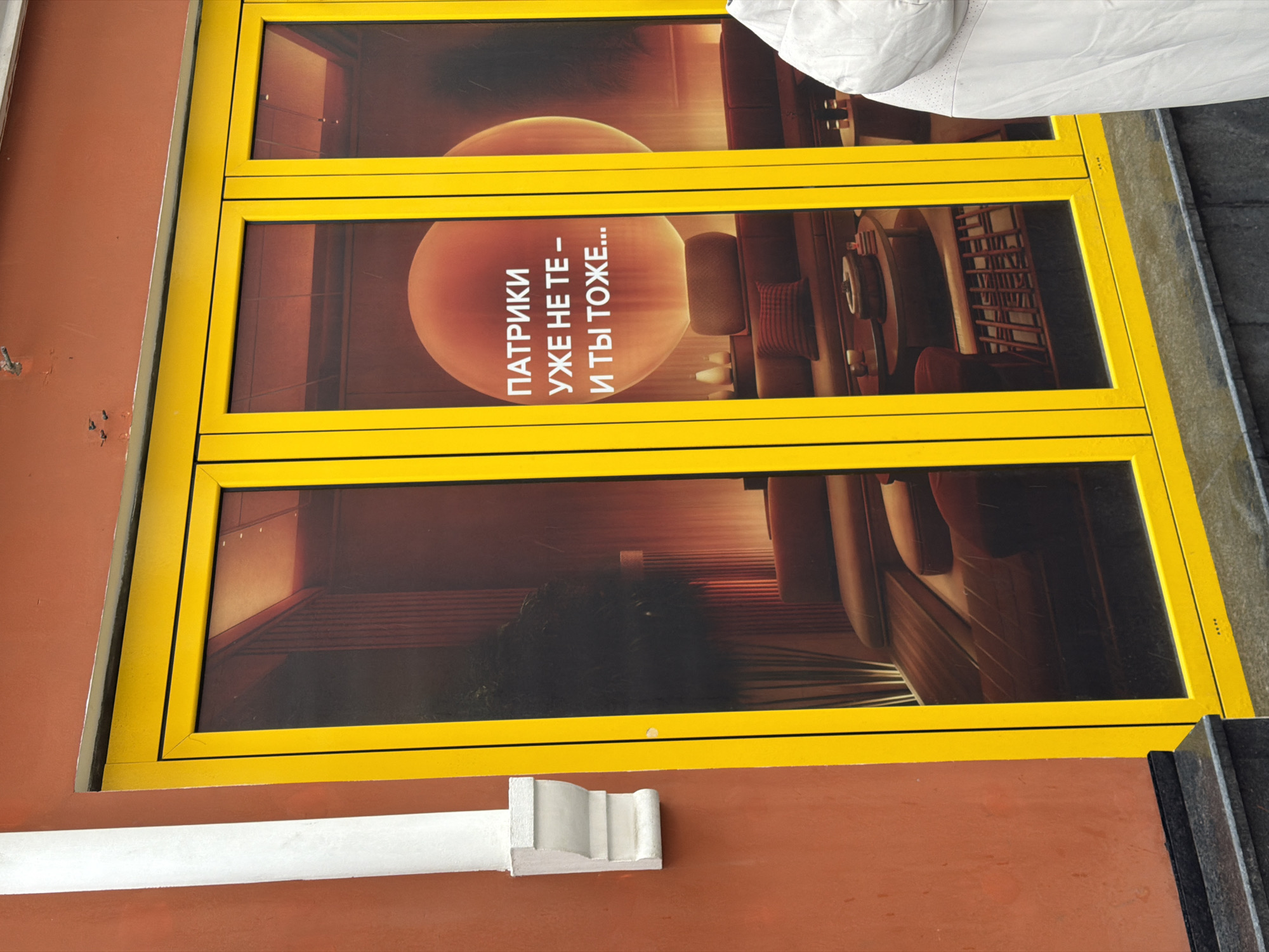 The image shows a close-up of a window display in what appears to be a commercial building in Moscow, Russia. The window frame is painted bright yellow, contrasting with the reddish-brown wall of the building. The window display features an advertisement poster in two parts. The upper part of the poster includes a blurry image of an interior space with an orange, glowing orb. Text in Russian is visible on the poster. The lower part of the poster continues the interior scene. A white architectural element (possibly a drain pipe) is located under the window.