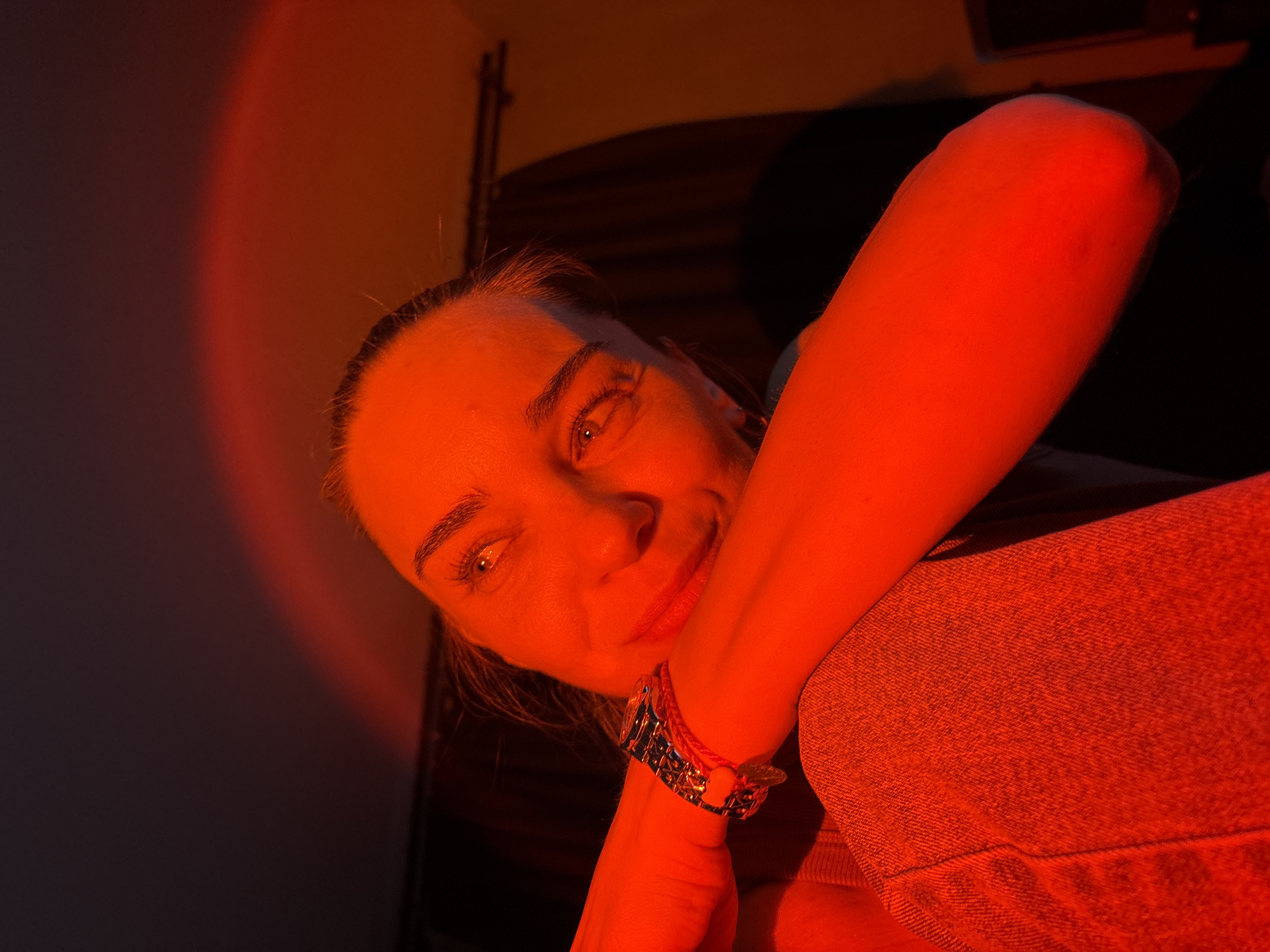 A close-up, low-angle shot captures a young woman bathed in intense red light. She is leaning on her arm, which is resting on her chest and shoulder, revealing a silver wristwatch. Her head is tilted back, and her gaze is directed upwards and to the left, with her eyes open and focused. The red lighting creates a dramatic effect, casting shadows and highlighting her features. The background is dark and out of focus, with hints of a curtain or drapes. The woman is wearing a denim garment, visible on her shoulder and arm. There is no indication of her location in the city of Unknown, Russia.