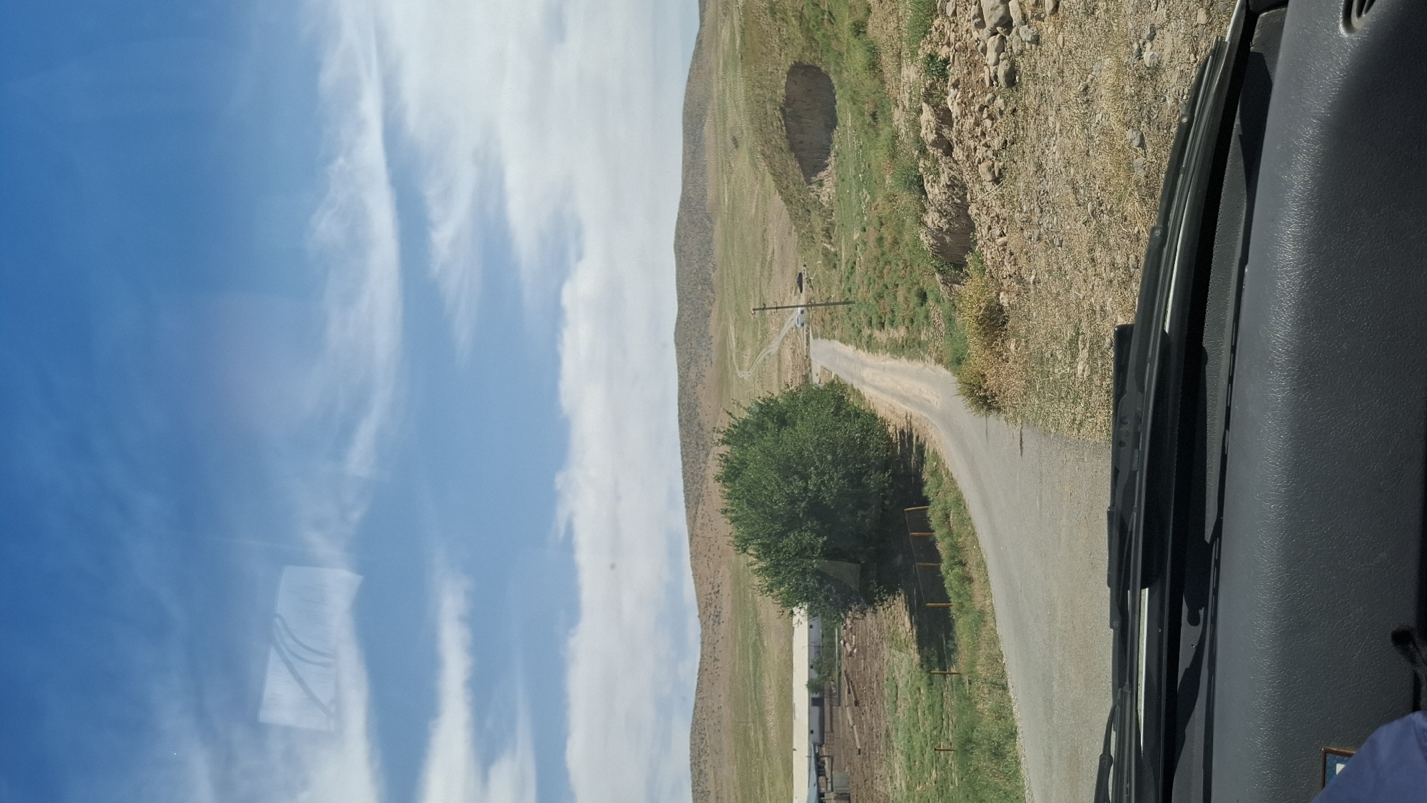 A paved road curves through a semi-arid landscape with rolling hills under a bright blue sky with scattered white clouds. To the left of the road, a large, dense green tree stands prominently. Further left, a long, low-lying building with a white roof is visible, suggesting a farm or industrial facility. The road ahead shows two vehicles, a white car and a dark-colored vehicle, parked on the right side. A utility pole stands near the parked vehicles. The terrain on the right side of the road is rocky and uneven, with a distinct depression or eroded area. The foreground shows the dashboard and windshield wipers of a vehicle, indicating the image was taken from inside a car. The location is identified as Boraldai, Kazakhstan, a city known for its dry, steppe climate and agricultural presence.