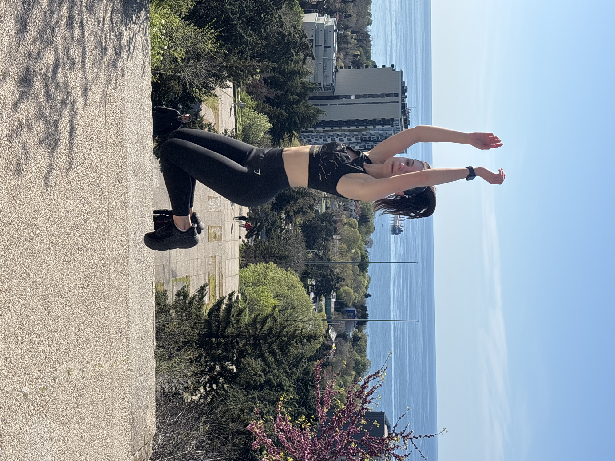 A young woman is captured outdoors, engaging in a fitness activity. She is dressed in black athletic wear, consisting of leggings and a patterned sports bra, and black athletic shoes. A dark smartwatch is visible on her left wrist. The woman is in a backbend or bridge-like pose, with her hips elevated and arms stretched upwards and backward, hands open. Her head is tilted back, and she is looking slightly upwards.
She is positioned on a light-colored, textured surface, possibly concrete or a paved area. The background reveals a scenic panorama. In the mid-ground, there are numerous green trees, some with lighter spring foliage, and several modern, multi-story buildings. Beyond the buildings and trees, a vast expanse of calm blue water, likely a sea or large lake, stretches towards the horizon under a clear, bright blue sky. A boat or ship can be seen on the water in the distance. In the foreground, a flowering tree with vibrant pink and purple blossoms adds a touch of color to the lower right of the frame.
The image appears to be taken on a sunny day, with clear skies, suggesting pleasant weather. The strong shadows indicate it is likely midday or early afternoon. A white logo, possibly "GYMSHARK," is visible on the outside of her left thigh.