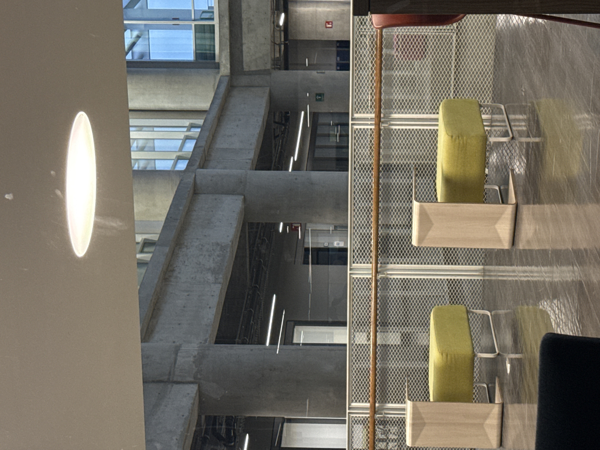 The image depicts the interior of a modern, multi-story building, oriented in a landscape format after being rotated from its original vertical capture. The scene is dominated by exposed concrete architecture, featuring thick pillars, beams, and interconnecting structures, suggesting an institutional or commercial building like an office, university, or public space.
In the foreground, separated by a light-colored metal mesh partition, are two modern seating units. Each unit consists of a vibrant lime-green cushioned top set into a light wooden or light-colored frame with a geometric design. A dark-colored chair or sofa section is partially visible in the bottom right corner. Reflections of the seating and the mesh are noticeable on a glossy floor or panel surface to the right of the seating. A wooden or metallic pole, possibly part of a floor lamp or stand, stands to the far right.
Beyond the mesh partition, the building's interior architecture unfolds across multiple levels. Large windows allow natural light to enter, reflecting a bright, possibly overcast, sky. Linear light fixtures or strips are visible on various ceilings and structural elements throughout the space. Exposed conduits or pipes run along some concrete surfaces, adding to an industrial-modern aesthetic.
Notable details include a red square sign with a white symbol (likely an emergency or safety indicator) on an upper concrete wall, and a green square sign with a white left-pointing arrow on a lower wall, indicating direction or an exit. No people are present, giving the impression of a quiet, unoccupied space. The strong natural light combined with artificial illumination suggests it is daytime.