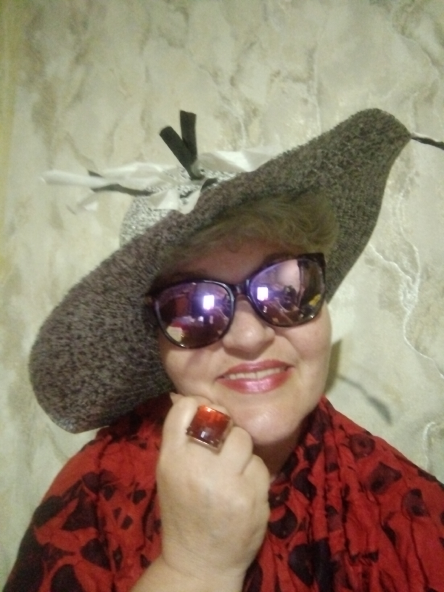 A woman wearing a wide-brimmed hat and large sunglasses poses for a selfie in front of a textured wall. She has a red, patterned scarf around her neck and is wearing a ring with a red stone on her right index finger. Her lips are painted with glossy pink lipstick, and she is smiling. The setting appears to be indoors, with the wall having a mottled, light beige and grey texture. The lighting is somewhat dim, causing reflections in her sunglasses. The image does not contain any specific landmarks or context to identify it as being in Horishni Plavni, Ukraine.