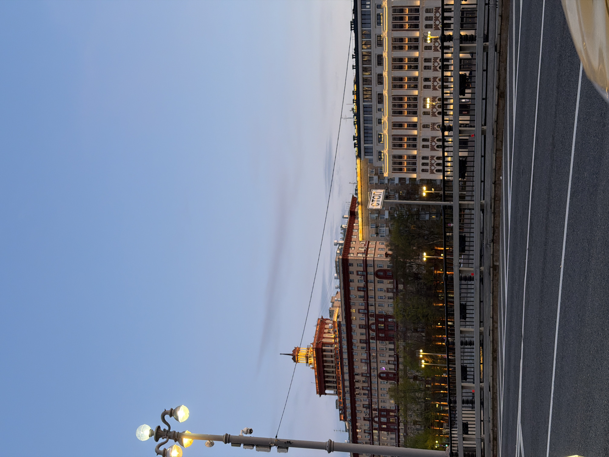 The image depicts a street in Moscow, Russia, with buildings lining one side and a road with white stripes on the other. Two multi-story buildings are visible, one with a red and white façade topped with a spire, and another with a white façade. Streetlights illuminate the buildings and road. A stop sign is affixed to a pole. The sky is light blue.