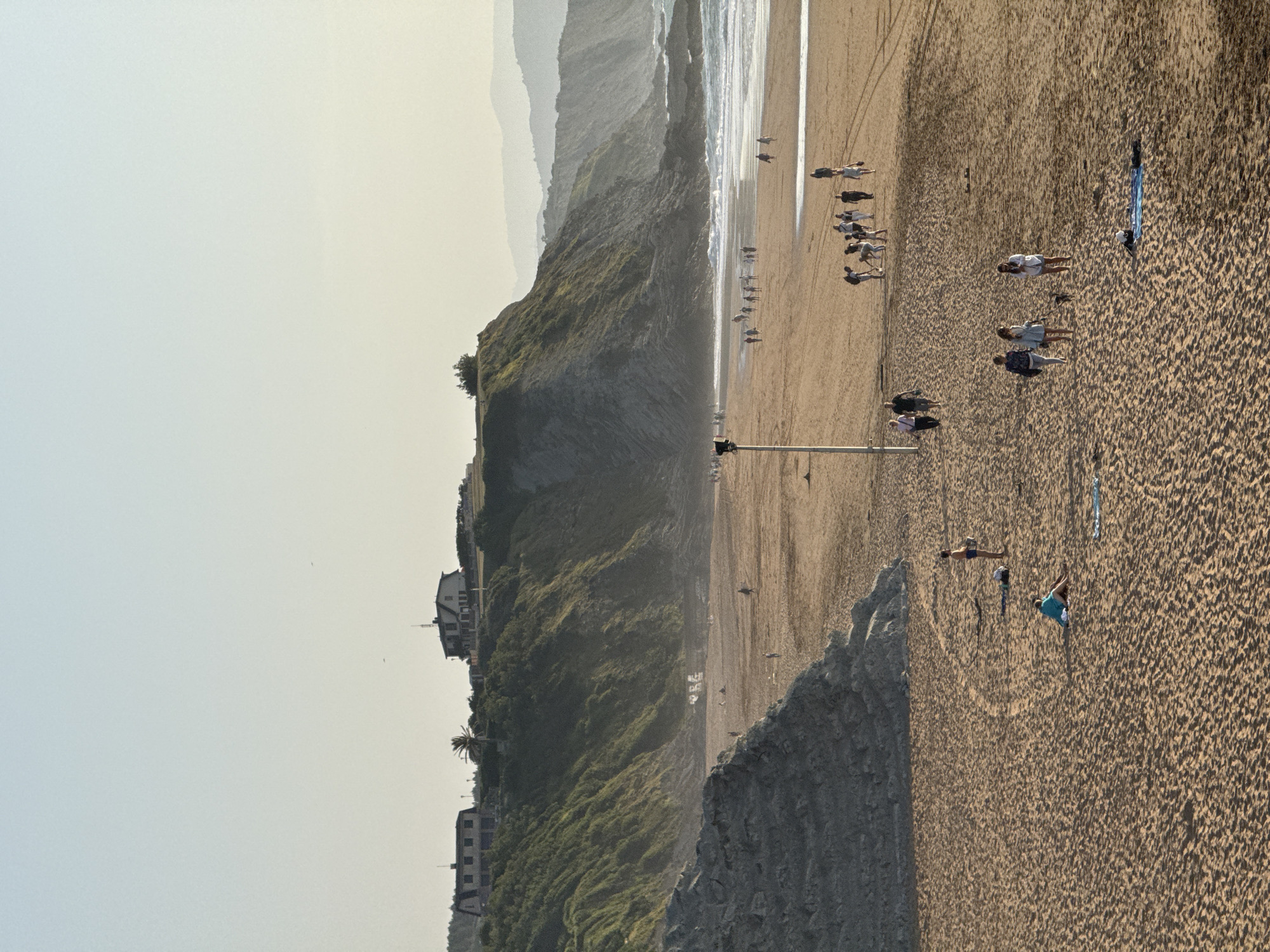 The image depicts a wide, sandy beach in Sopela, Spain, at golden hour. A gentle ocean surf breaks on the right side of the frame, meeting a rocky shoreline. In the foreground, people are scattered across the sand. Some are walking, while others are sitting or lying on towels. A group of individuals is clustered together, appearing to be engaged in an activity, possibly playing. Further back on the beach, more people are walking or standing. To the left, a large, gray rock formation dominates the scene. In the middle ground, a light pole stands tall. In the background, a lush, green cliff rises, topped by two modern buildings. The sky is clear and pale, with a hazy, mountainous landscape visible in the far distance. The overall impression is one of a peaceful late afternoon at the seaside.