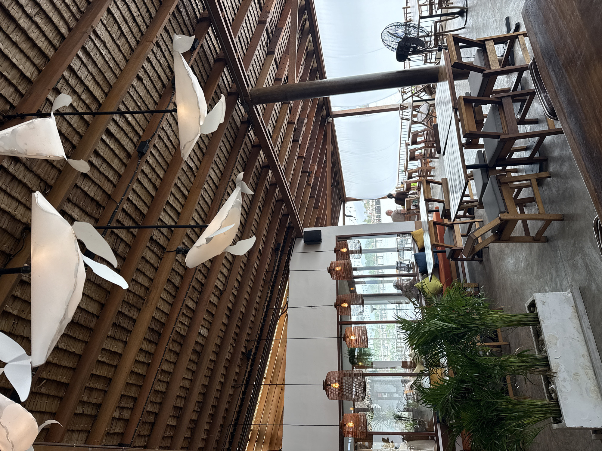 This is a medium-wide, eye-level shot of an open-air restaurant, likely located in the Ko Samui District of Thailand, given the tropical atmosphere and design elements. The ceiling is constructed from a thatched roof supported by wooden beams. Suspended from the ceiling are several large, white, whale-shaped light fixtures. Below, the restaurant is furnished with wooden tables and chairs. In the foreground, a planter box contains several lush green palm plants. Large windows or open doorways offer a view of the outdoors, possibly a waterfront, with distant hills visible. A large industrial fan is present on the right side of the image. Several patrons are seated at tables, engaged in dining or conversation. The overall ambiance suggests a casual dining establishment with a nautical theme.