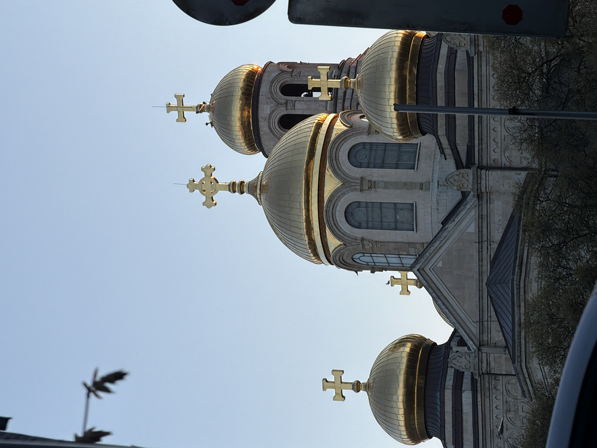 The image shows the domes of the Dormition of the Theotokos Cathedral in Varna, Bulgaria, viewed from below. The domes are gilded and topped with crosses. The sky is light blue. Architectural details of the cathedral's facade are visible. There is foliage from trees on the right side of the image. Part of a vehicle is visible at the bottom and top of the image, suggesting the photo was taken from within a car.
