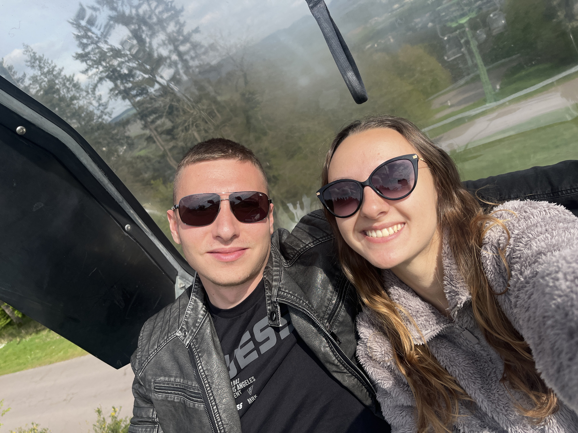 A selfie shows a Caucasian male and female inside a cable car with a clear dome. The man, left, is wearing a black Guess t-shirt and a gray leather jacket, along with sunglasses. The woman, right, is wearing a gray fluffy sweater and sunglasses. Behind them, seen through the cable car dome, are green trees and buildings. The location is in Saarburg, Germany, as evidenced by the cable car system, a tourist attraction in the area. They are both smiling.