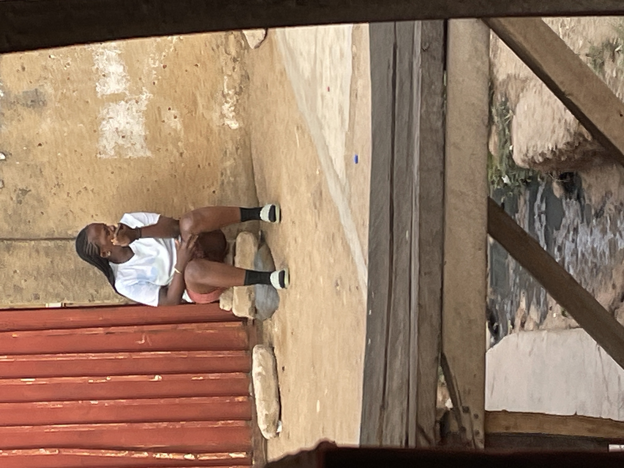 A woman with braided hair is sitting outdoors, eating something she holds in her hand. She is wearing a white t-shirt, dark socks, and light-colored shorts. She sits on a concrete or stone step or ledge. The location appears to be Jos, Nigeria. To the left, a red corrugated metal wall is visible. Beside her, is a rough, beige concrete or dirt ground surface. To the right, there is a structure constructed of wooden planks and beams. Beyond this structure, there is a rocky and vegetated terrain.