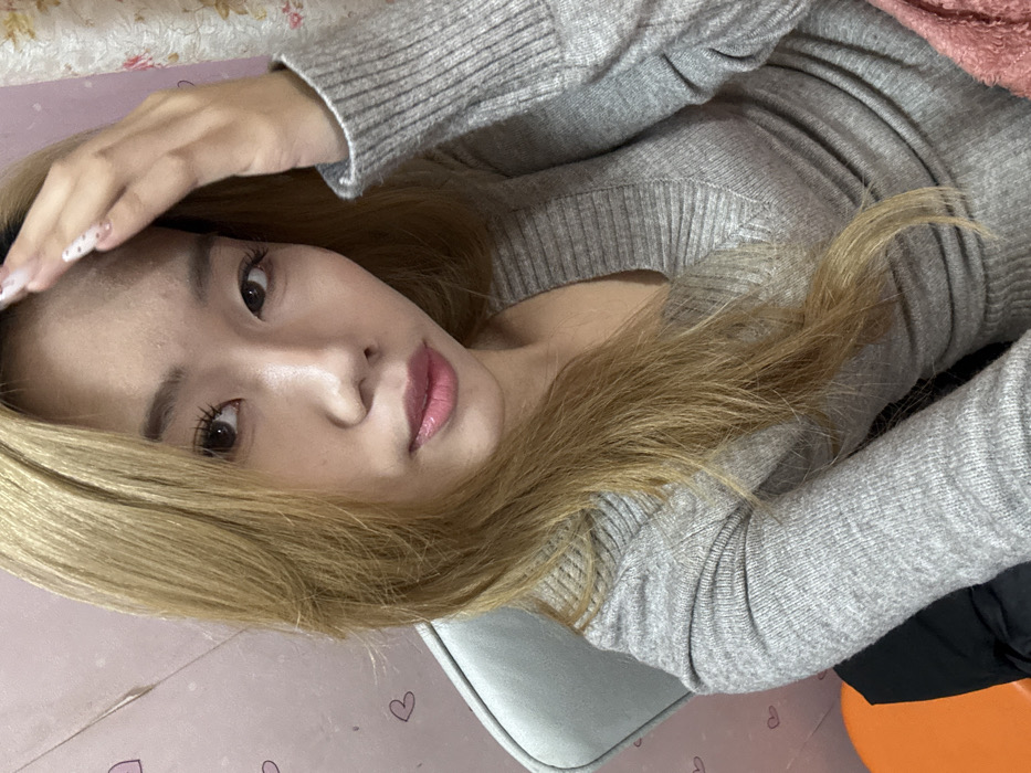 A young woman with light brown hair and fair skin poses for a photo. She wears a ribbed, long-sleeved gray shirt and has long, manicured nails. Her head is tilted, and she gazes into the camera with a subtle smile, her hand near her forehead. The background includes a light pink surface with heart decorations and what appears to be a cushion or chair.