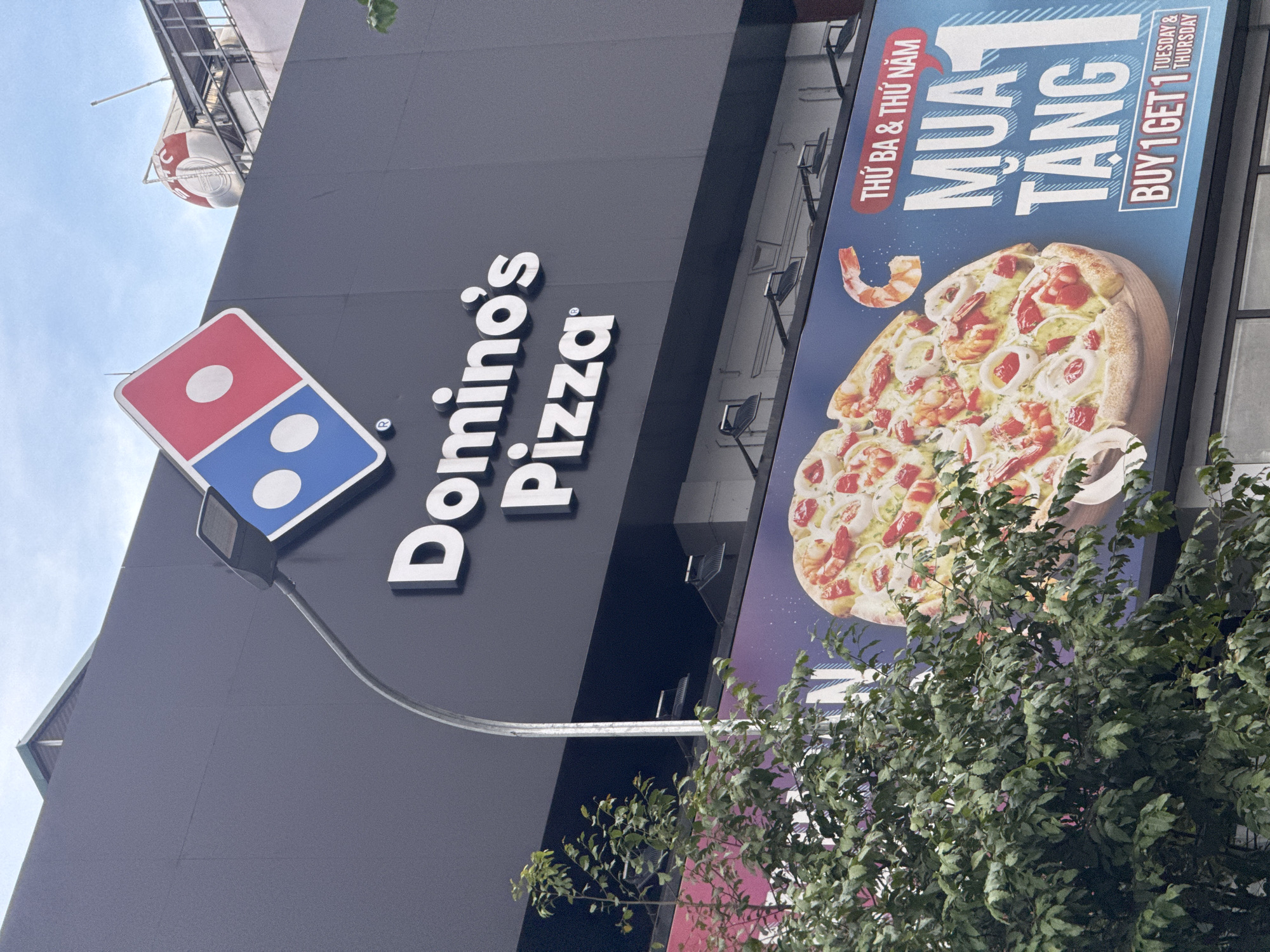 The image shows the Domino's Pizza signage and a billboard advertisement outside a building in Hanoi, Vietnam. The Domino's logo, a red and blue diamond with white dots, is prominently displayed on the dark grey facade of the building. Below the logo, the words "Domino's Pizza" are written in large white letters.
A billboard below the signage features a large image of a pizza with shrimp and calamari, alongside text in Vietnamese and English. The Vietnamese text "THỨ BA & THỨ NĂM MUA 1 TẶNG 1" translates to "Tuesday & Thursday Buy 1 Get 1". The English text below confirms this promotion: "BUY 1 GET 1 TUESDAY & THURSDAY".
A street lamp extends from the building, casting light downwards. In the foreground, a leafy green tree partially obscures the view of the lower part of the building and the billboard. The sky above is blue with some clouds. The building appears to be a commercial establishment, likely in an urban area of Hanoi.