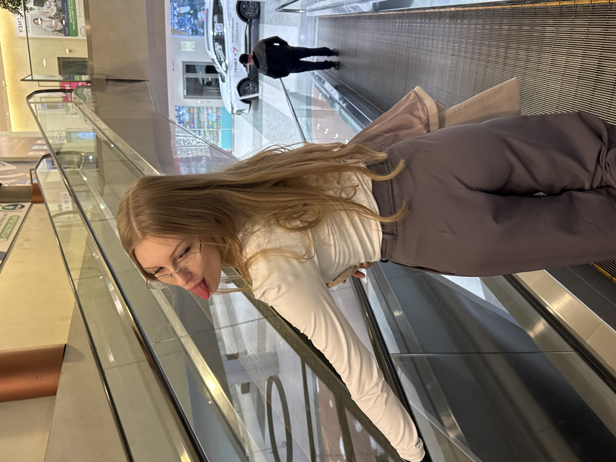 A fair-skinned woman with blonde hair and glasses is standing on an escalator indoors in Perm, Russia. She is sticking her tongue out. She is wearing a long-sleeved white shirt and brown pants. A person in a black jacket is standing on a parallel escalator. A white car is visible in the background. There are signs and advertisements on the wall.