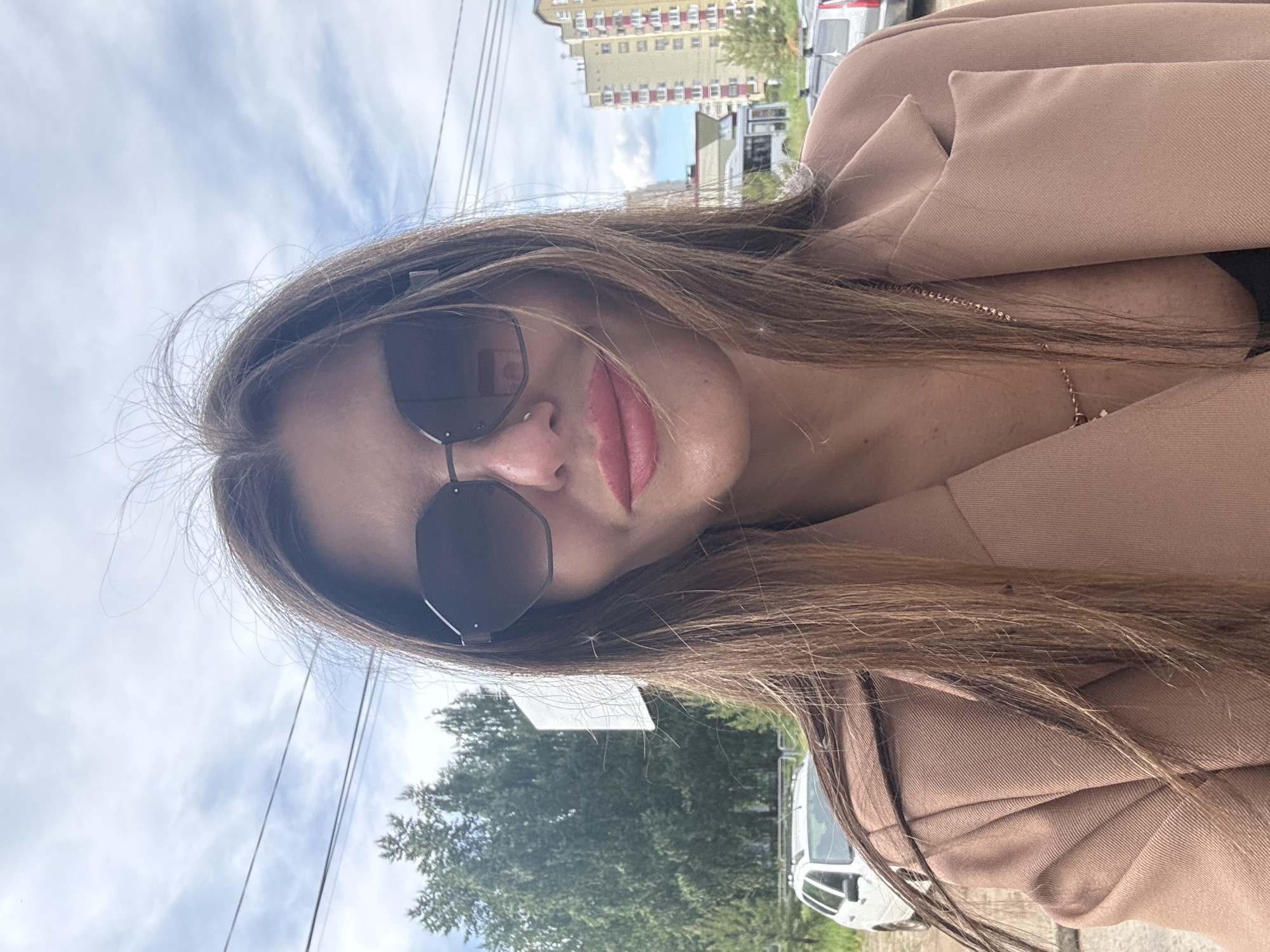 This image is a rotated, close-up selfie-style portrait of a woman. The image is oriented with the woman's head tilted significantly, making it appear as if she is looking upwards. She has long, straight light brown hair, parted on the side, framing her face and falling over her shoulders. She wears dark, octagonal-shaped sunglasses that obscure her eyes. Her lips are painted with a soft pink or nude lipstick, showing a slight, subtle smile.
She is dressed in a light brown or beige blazer, under which a dark top and a thin golden chain necklace are visible around her neck.
The background shows an outdoor setting on what appears to be a bright, partly cloudy day. Above her, against the sky, are several utility lines. In the distance, typical residential buildings, possibly apartments of Soviet-era architecture, can be seen, with one prominent light yellow building. Green foliage from trees is visible, suggesting a street or park area. Parts of several vehicles, including a white van or car, are also visible towards the lower and right edges of the frame.
The overall impression is a casual, daytime self-portrait taken in an urban or suburban environment in Chaikovsky, Russia. No text is visible in the image.