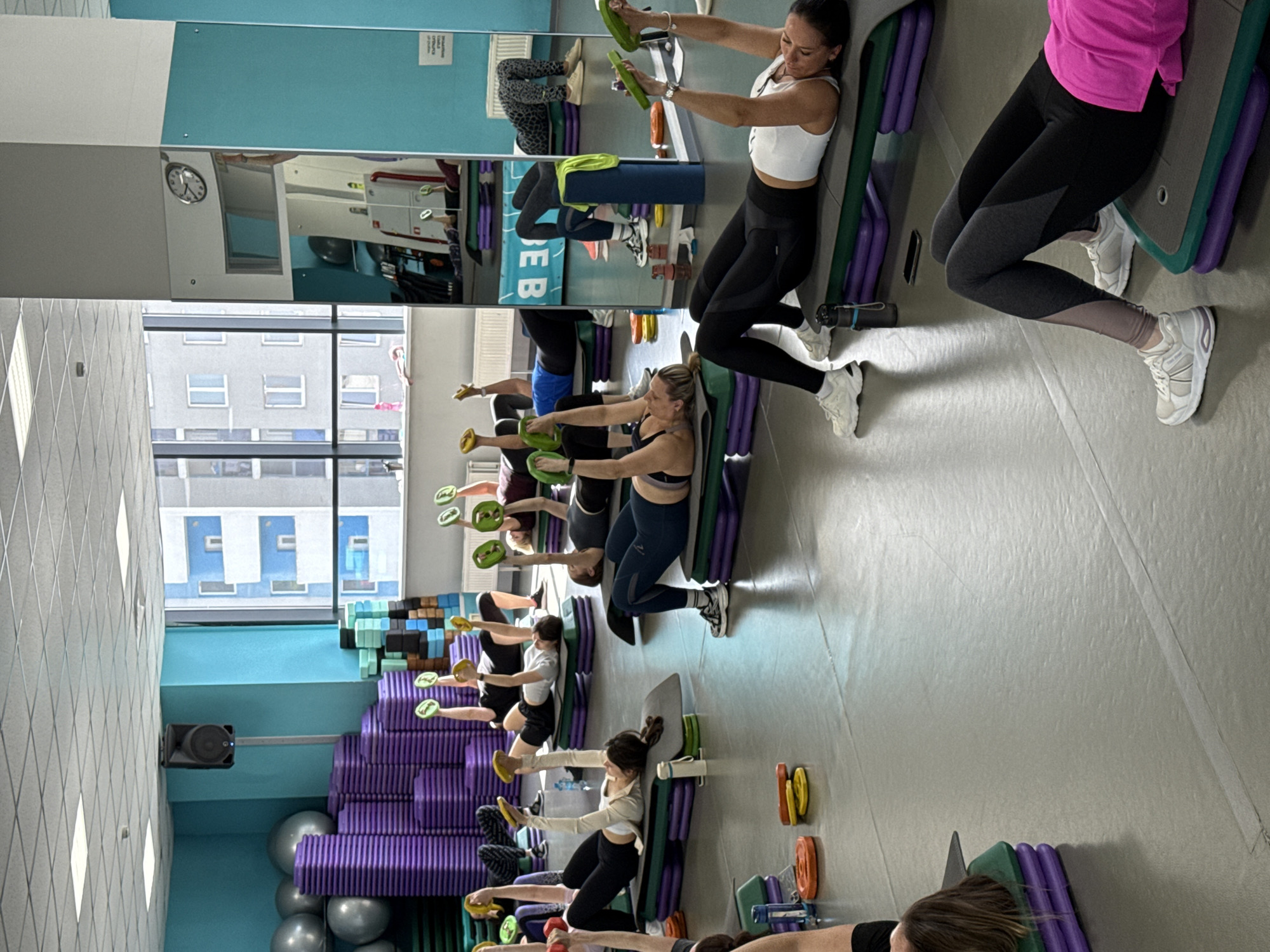 In an indoor fitness studio in Yekaterinburg, Russia, several women are engaged in a group exercise class. The women are dressed in athletic wear, including leggings and sports tops, and are performing exercises, many holding small, disc-shaped weights in green or yellow. They are positioned along a wall, using exercise steps or benches in purple and green for support, suggesting a core or resistance training workout. Their expressions indicate focus and engagement.
The studio features light-colored walls, a light gray floor, and accents of bright blue on the upper walls. A large mirrored wall reflects parts of the room, including participants and equipment, and distorts some elements. In the reflection, a wall clock shows the time to be approximately 12:45. Another reflection shows text that appears to be "ВНИМАНИЕ!" (Attention!) on a sign. Large windows line one side of the room, offering a view of a modern, multi-story urban building with numerous windows outside, indicating a daytime setting.
Various gym equipment is visible, including stacks of purple foam rollers or mats, several gray exercise balls, and a black speaker mounted on a blue wall section. Personal items like water bottles are placed near some participants. The overall atmosphere is that of a well-equipped, bright, and active fitness environment.