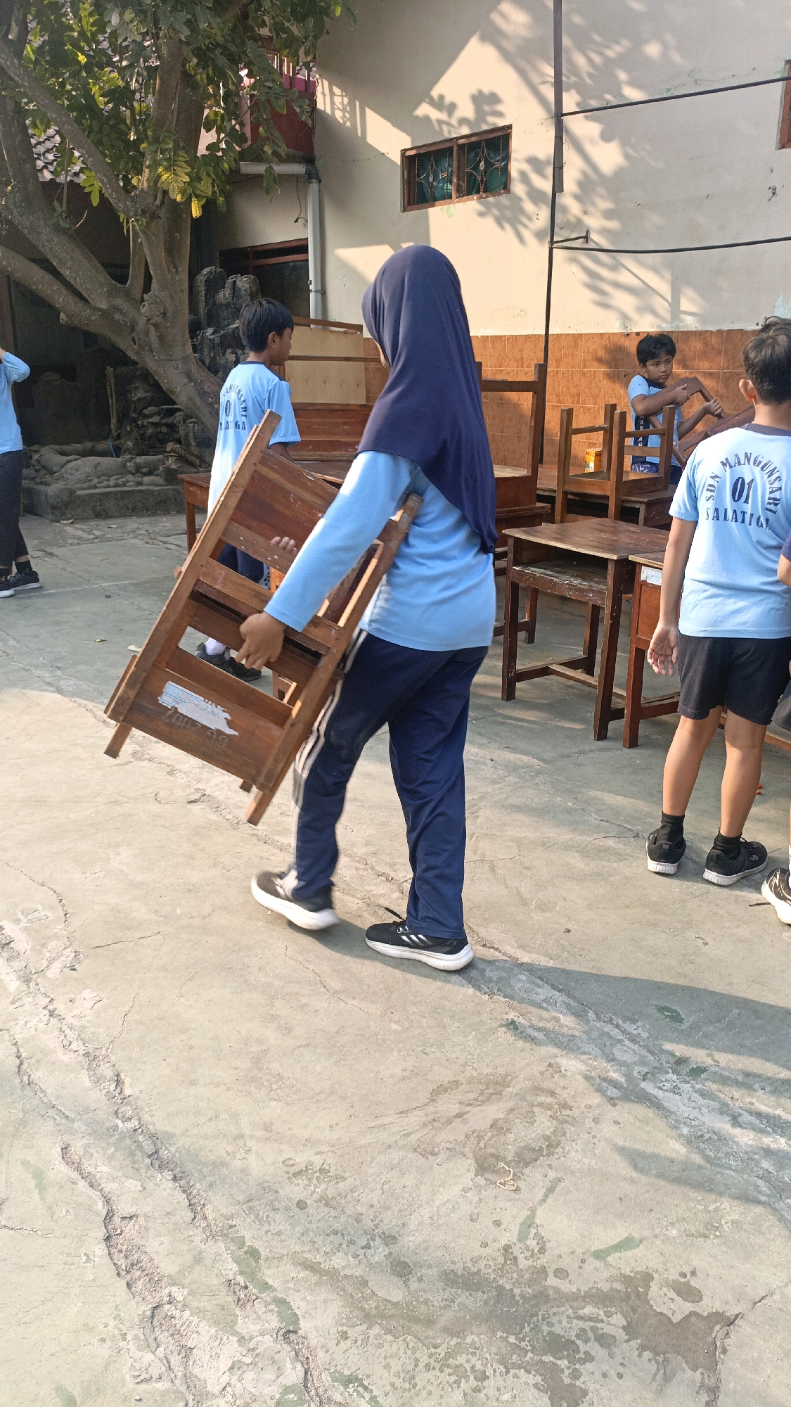 In the city of Salatiga, Indonesia, a student wearing a navy blue hijab and a light blue long-sleeved shirt and navy blue track pants carries a wooden chair with a partially visible white label on its side. The student is walking on a cracked concrete surface. Behind the student, other students in light blue shirts are visible, along with wooden desks and chairs, suggesting an outdoor school setting, possibly a playground or courtyard. The sunlight casts shadows on the ground. The background includes a building with tiled walls and windows, and a large tree with leafy branches.