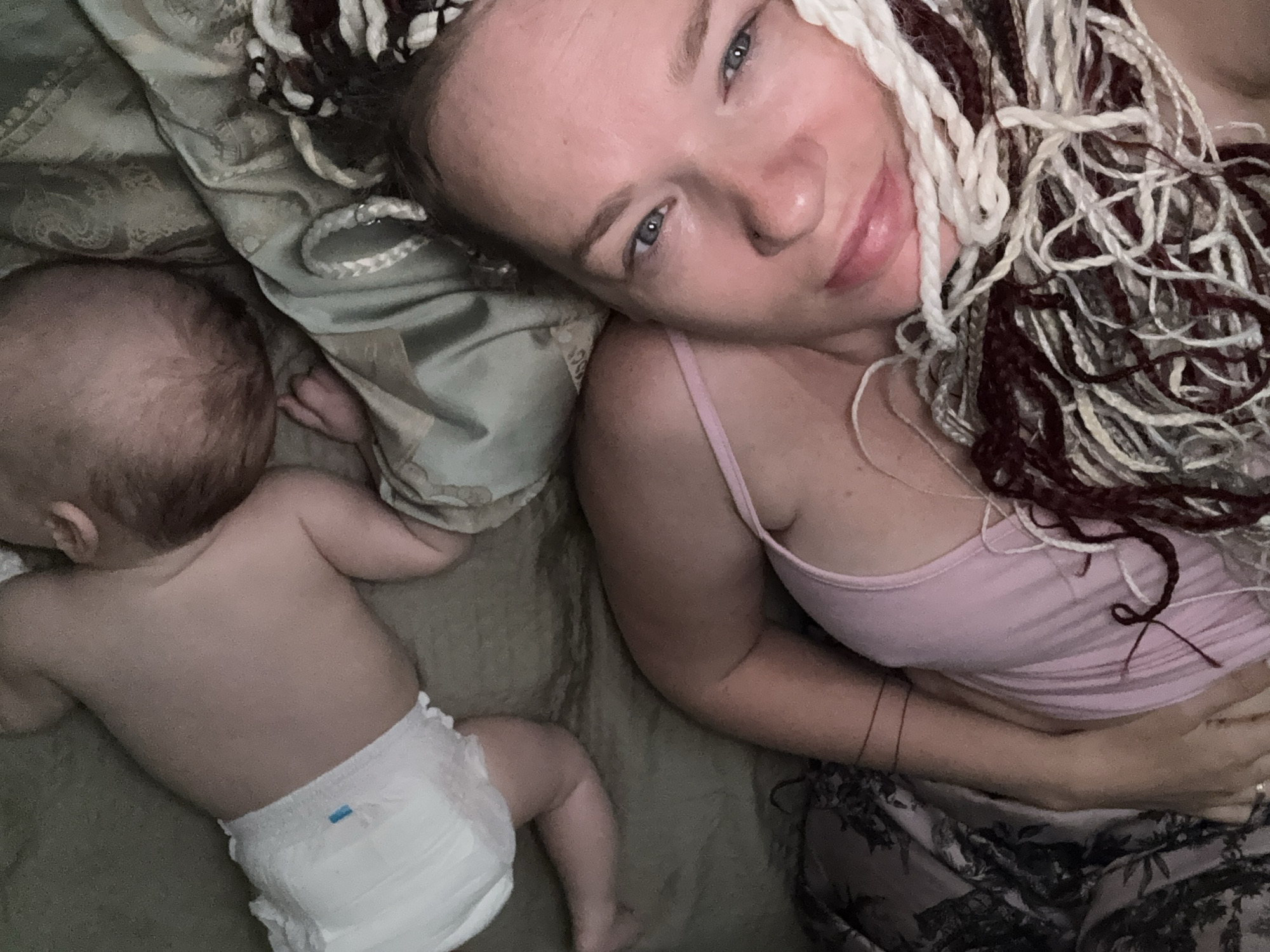 The image shows an overhead, close-up view of a woman and a baby lying on a bed. The woman is in the foreground, facing the camera, with her eyes open and looking towards it. She has blue eyes and light-colored skin. Her hair is styled in long braids with streaks of white and dark red. She is wearing a pink tank top. The baby is in the background, lying on its stomach and facing away from the camera. The baby is wearing a white diaper. The bed has a patterned duvet in muted colors. The scene appears to be indoors, possibly in a bedroom. There is no indication of the location being in Moscow, Russia.