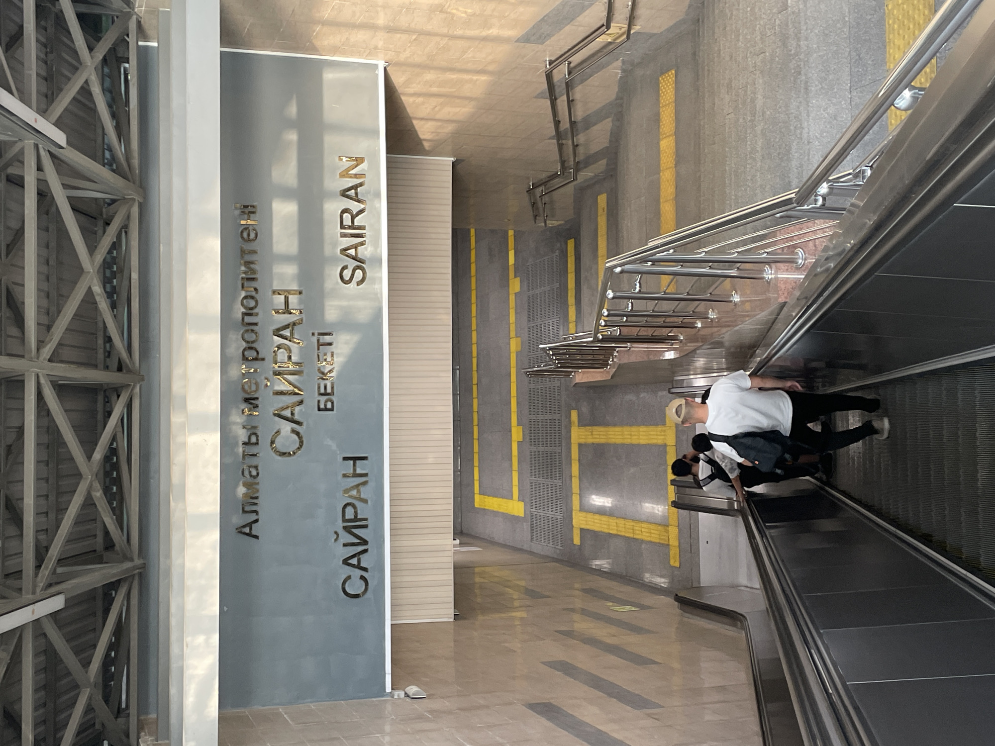 An upward-facing, full shot captures the entrance of the Sairan metro station in Almaty, Kazakhstan. The station name is prominently displayed in large, metallic letters on a grey, rectangular sign. Above the station name, smaller metallic letters spell out "Алматы метрополитені," which translates to "Almaty Metro." Below the station name, more metallic letters read "SAIRAN" in larger font, with "БЕКЕТІ" in smaller font centered above it.
The scene shows a downward-sloping escalator on the left and a set of stairs with metal railings on the right, both leading away from the viewer. Yellow tactile paving strips are visible on both the escalator and stair landings, indicating accessibility features. Three individuals are partially visible on the escalator. The person closest to the camera is a male wearing a white t-shirt, black pants, and a beige baseball cap, carrying a black backpack. The two individuals ahead of him are dressed in white t-shirts and dark pants, also carrying backpacks. The walls are clad in a light-colored tiled material, with darker rectangular accents on the left wall. The ceiling structure, made of grey metal beams, is visible at the top of the image. The overall lighting suggests daylight, with some shadows indicating overhead lighting or natural light sources.