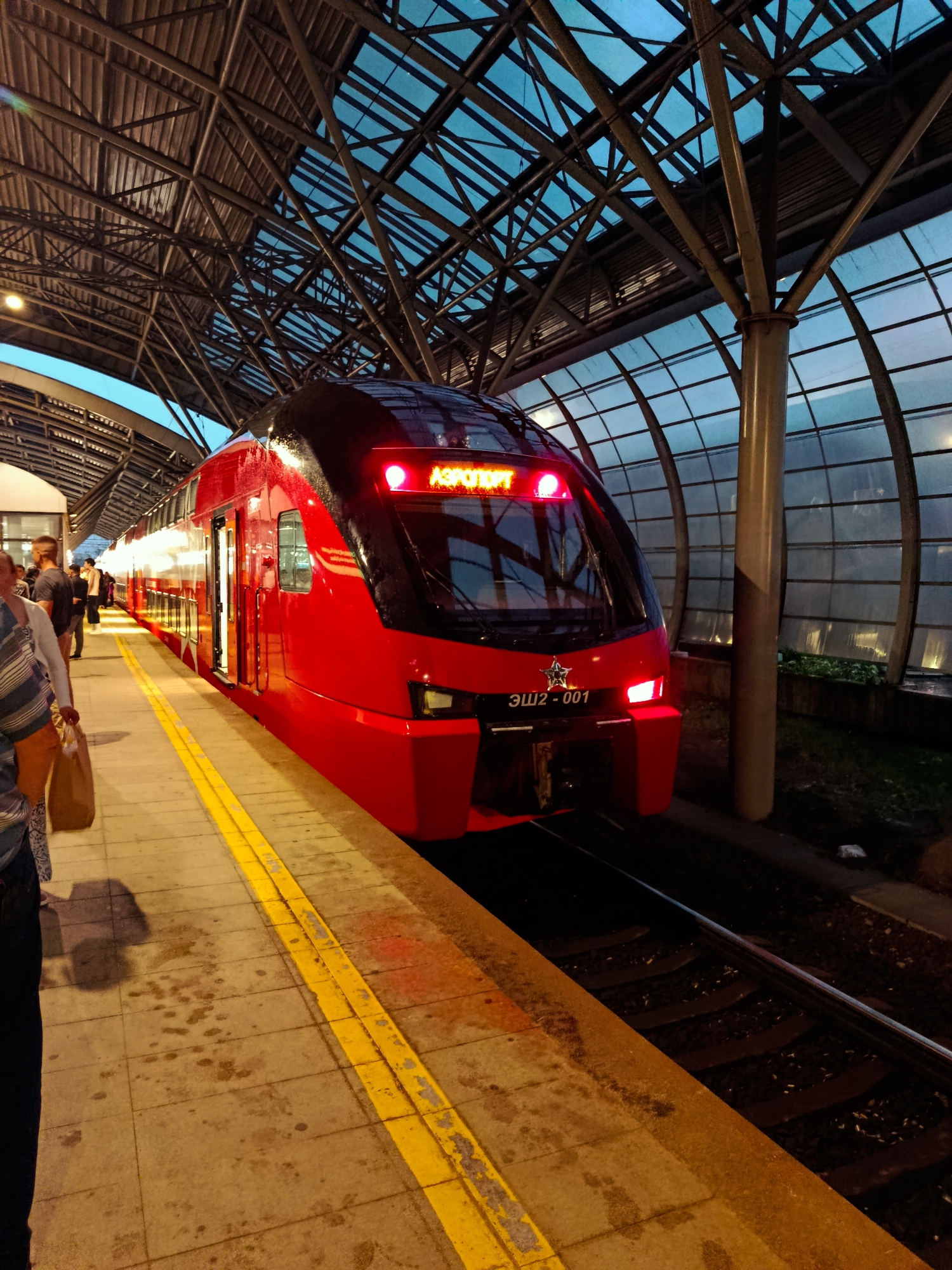 The image depicts a red double-decker train at a station in Moscow, Russia. The train, identified with the number ЭШ2-001, is modern and sleek, with its headlights illuminated. The station features a distinctive architectural design with a large, arched, glass-covered roof supported by metal beams, allowing for natural light to filter in. The platform is paved with concrete tiles and has a yellow tactile paving strip along the edge. Several people are visible on the platform, some standing and others walking, suggesting it is a public transport hub. The overall lighting suggests it is either early morning or late evening, with the artificial lights of the station also contributing to the illumination. The presence of the train and the station infrastructure points to this being a railway station, likely serving a commuter or express route, possibly to or from an airport given the word "Аэропорт" visible on the train's display. The environment appears clean and well-maintained.