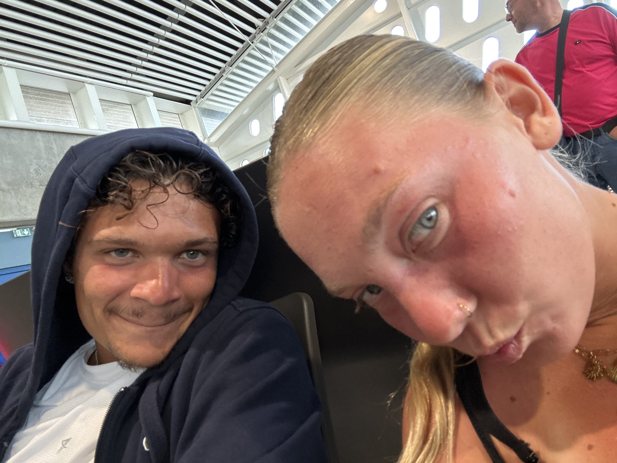 Two individuals are featured prominently in a selfie-style photograph. On the left, a young male with curly brown hair is wearing a navy blue hooded sweatshirt over a white t-shirt. He has light eyes and a mustache, and is smiling slightly. To the right, a young female with blonde hair pulled back, is seen making a pouting expression with her lips. She has fair skin, blue eyes, and a nose piercing. A black garment is visible on her upper body.
Behind the two, a portion of a building interior is visible. A section of a ceiling or roof structure with parallel white beams is present. In the upper right corner, a blurry figure of a man in a red shirt with a dark shoulder bag is partially visible.
The image was taken inside an unidentified building in Les Abymes, France.