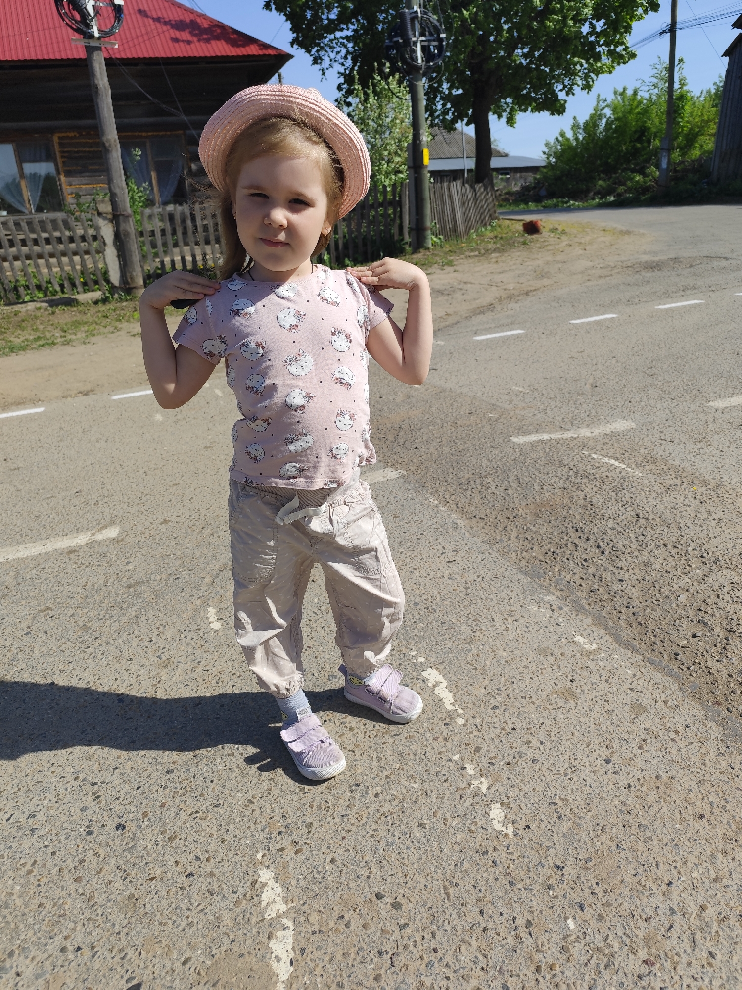 A young girl stands on a paved road with broken white lines. She is wearing a pink shirt with cat designs, light-colored pants, a pink hat, and pink shoes. Her hands are resting on her shoulders. In the background, there are wooden houses, a wooden fence, trees, and a utility pole. The image was taken in Фоки, Russia.