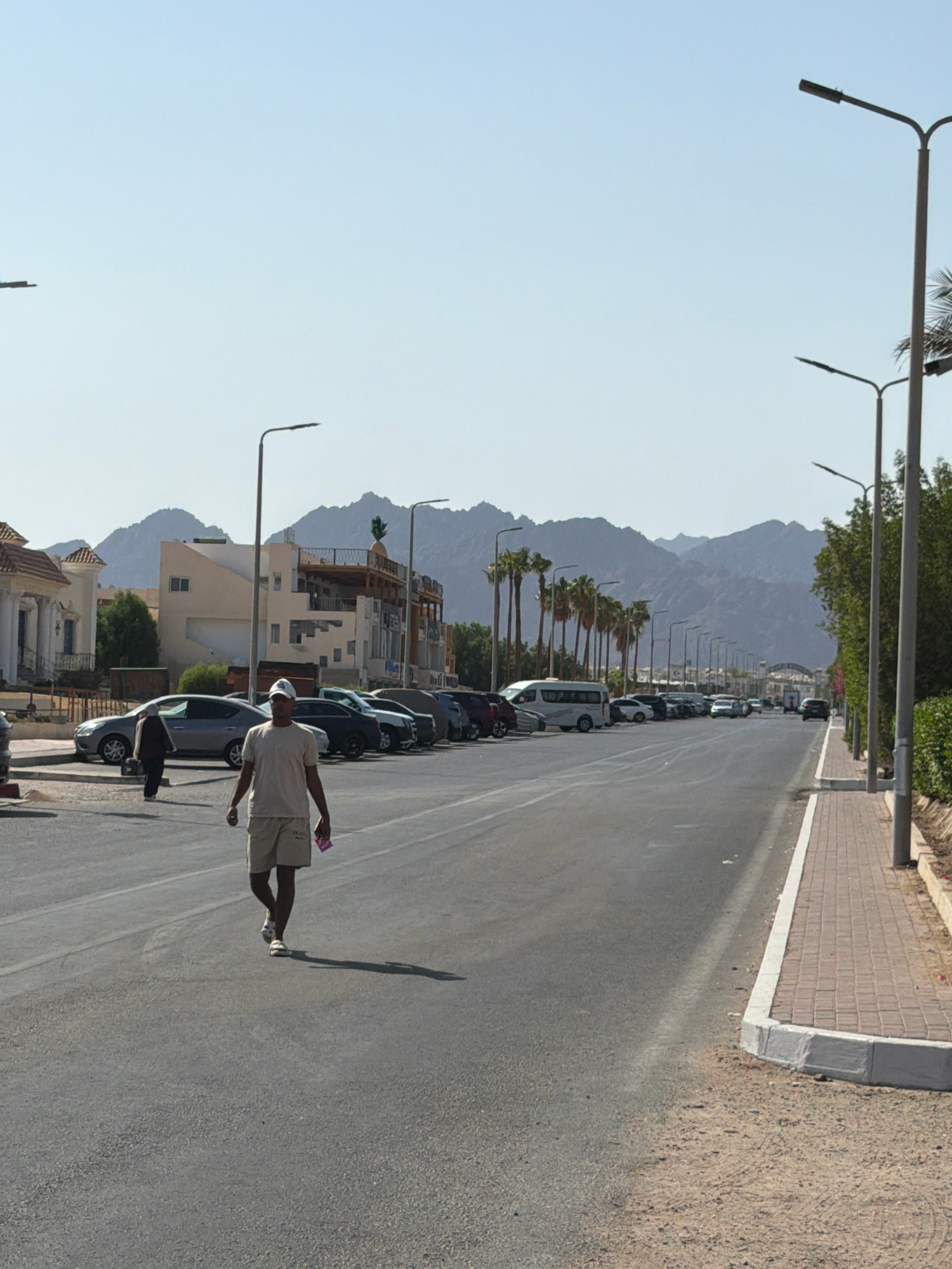 The image depicts a street scene in Sharm El Sheikh, Egypt, characterized by arid surroundings and mountainous terrain in the background. A man is walking towards the camera on the left side of the road, wearing a light-colored t-shirt, shorts, and a cap. He is holding a small object in his right hand. A line of parked cars and vans is visible along the left side of the road. On the right side of the road, a paved sidewalk with brickwork runs parallel to the street, bordered by a sand and gravel shoulder. Several streetlights are positioned along the sidewalk. In the distance, buildings with distinctive architecture and palm trees line the street, leading towards a more distant, hazy mountainous landscape. The sky is clear and blue, indicating a sunny day.