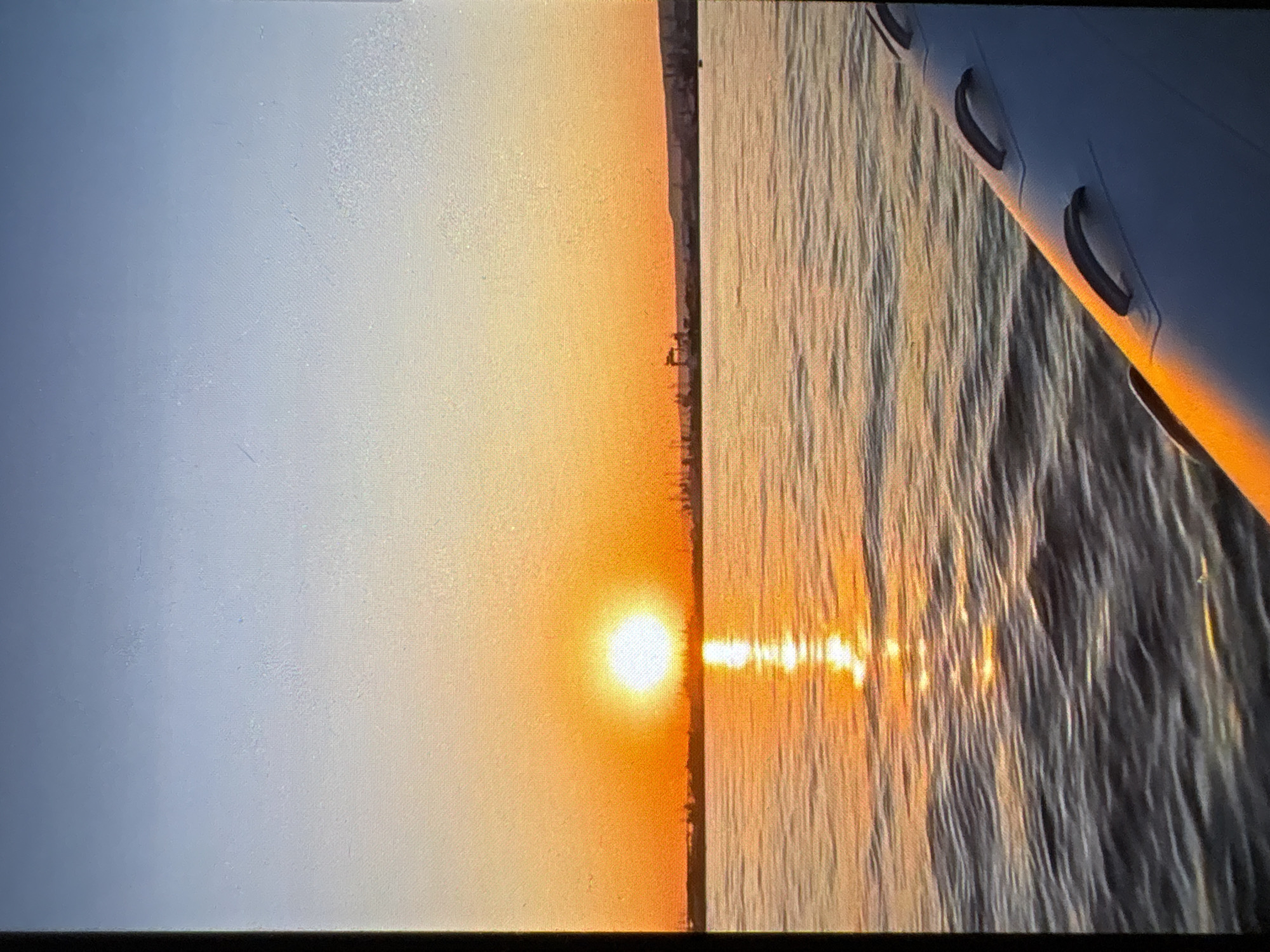 The image depicts a sunset over water. The sun is positioned on the horizon, its reflection shimmering on the water's surface. A coastline is barely visible as a dark line between the sky and water. The wing of an aircraft is visible in the foreground, indicating the photograph was taken from aboard a plane. There is no additional information to identify the city.