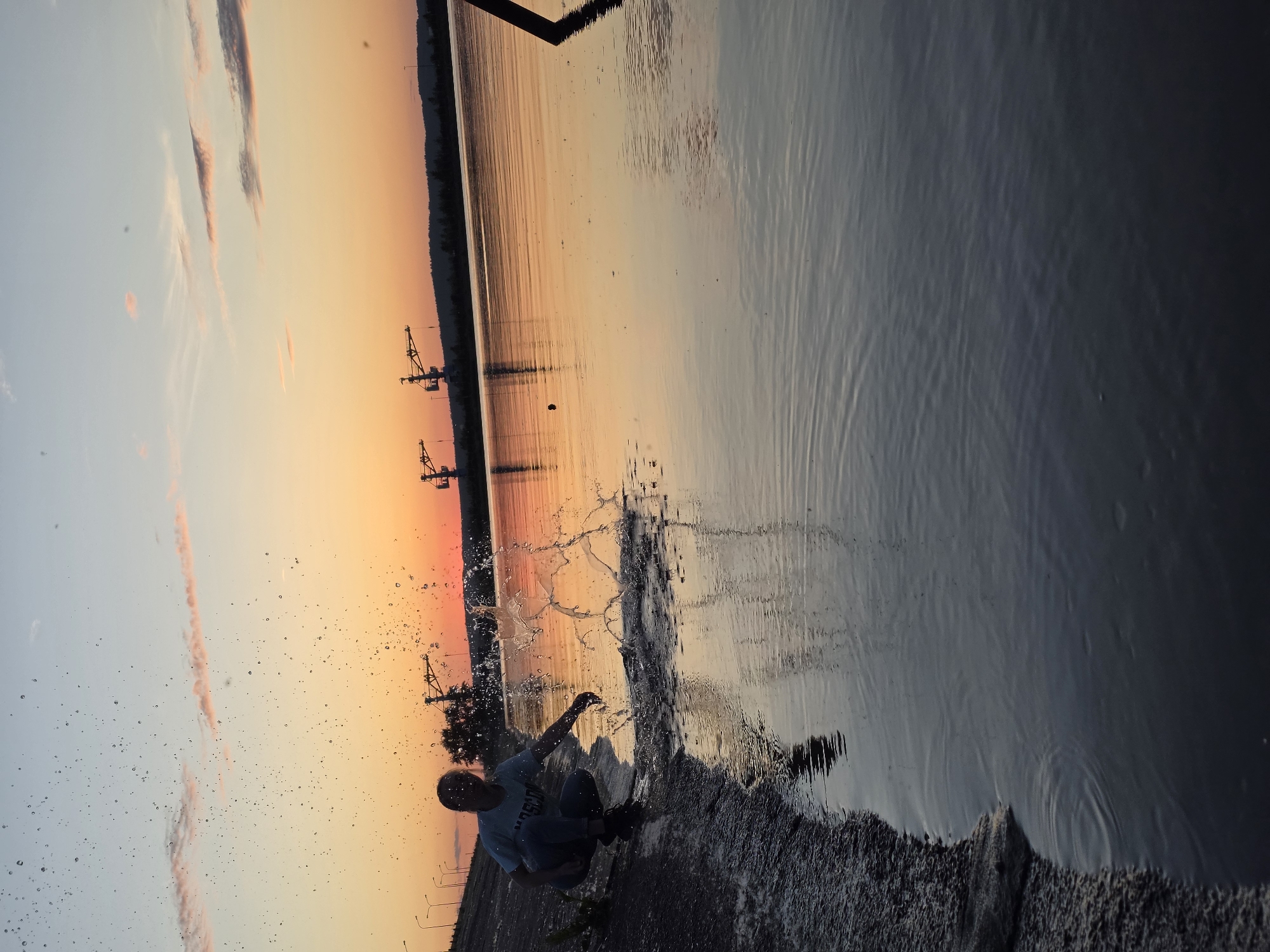 This image depicts a sunset scene in what is identified as Chaikovsky, Russia. A person wearing a light-colored t-shirt with visible text and dark trousers is captured mid-action, squatting on a rocky embankment at the edge of a calm body of water. Their right hand is immersed in the water, generating a significant splash with numerous droplets suspended in the air, illuminated against the bright horizon. The sky exhibits a gradient from pale blue at the top to a vibrant orange and red near the horizon, indicative of sunset, with scattered light clouds. The water reflects these warm sky colors. In the mid-ground, a dark treeline and several large industrial cranes are silhouetted against the colorful sky. A row of streetlights lines an elevated structure or road on the left, and a dark vertical post is partially visible on the far right of the frame.