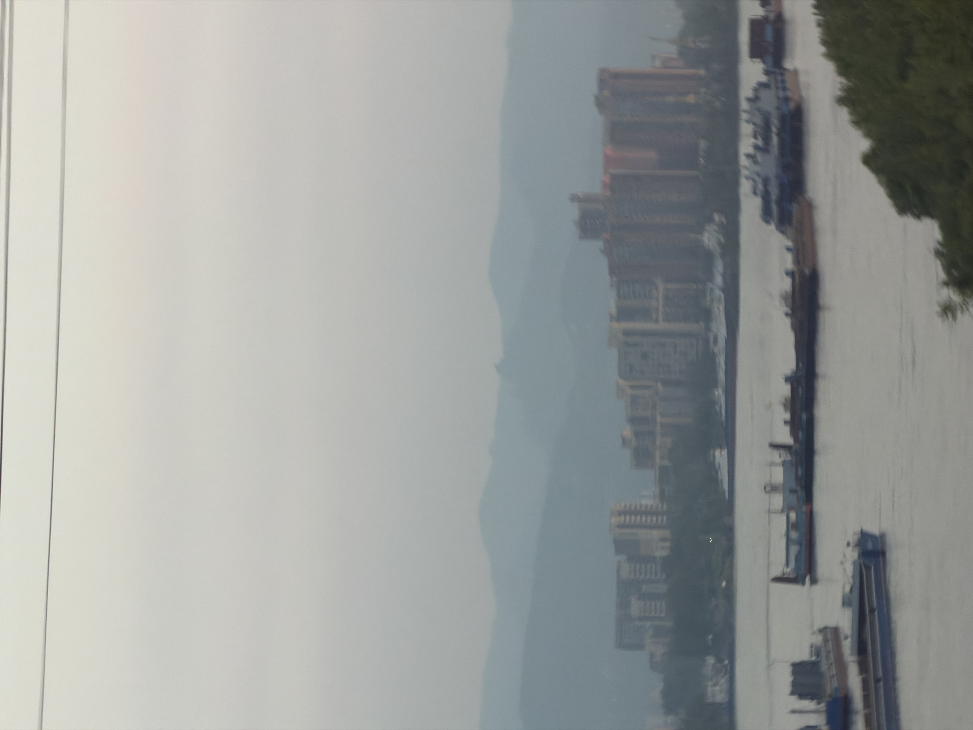 The image shows a hazy, overcast day in Krasnoyarsk, Russia, with a wide river in the foreground and a distant cityscape with hills behind it. Several barges and boats are on the river, some appearing to be stationary and others potentially in motion. The buildings in the distance suggest a developed urban area, with some high-rise structures visible. The overall atmosphere is subdued due to the weather conditions, with muted colors and soft focus throughout the scene. The image likely captures a view of the Yenisei River and the city of Krasnoyarsk from a vantage point overlooking the water.