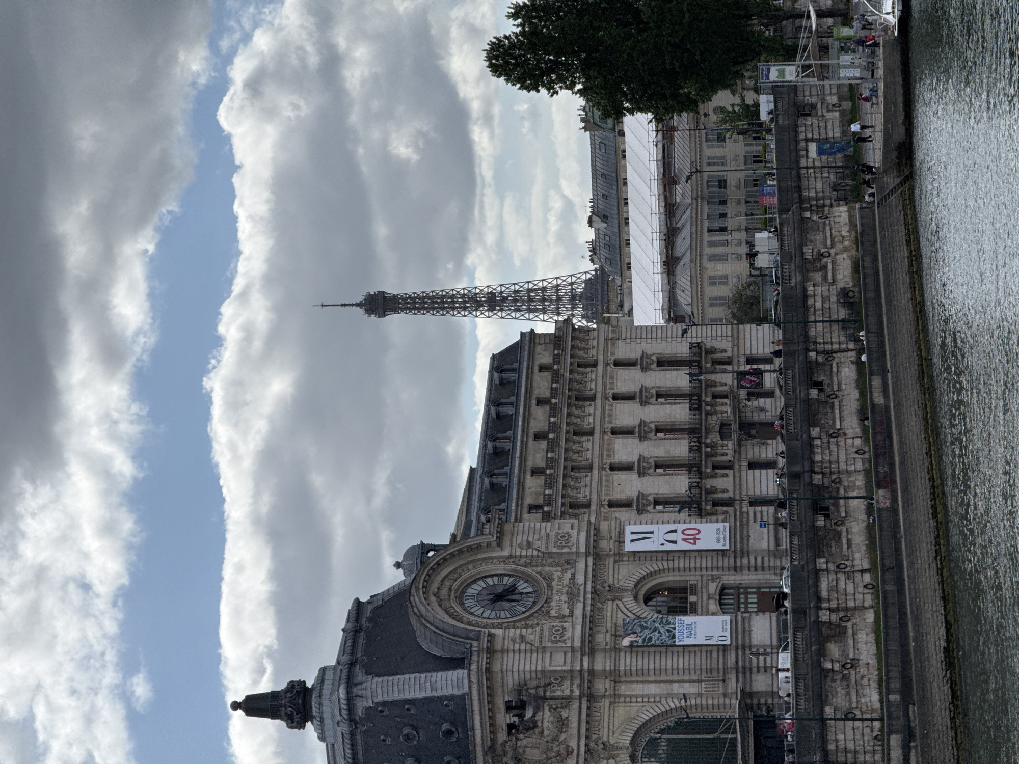 This image presents a picturesque urban scene in Paris, France, showcasing iconic landmarks along the Seine River. The foreground is dominated by the grand, ornate stone facade of the Musée d'Orsay, featuring its distinctive large clock face with Roman numerals. Above the clock, text reads "RO" and "PARIS ORLEANS," hinting at its history. Two prominent banners adorn the building: one displays "M 40" in red, with "1986-2026 Musée d'Orsay" below, indicating a 40-year anniversary celebration. Another banner advertises "YOUSSEF NABIL" and "LA REVANCHE DU MUSEE," likely for an exhibition.
To the right of the museum, the serene waters of the Seine River flow, its surface marked by gentle ripples. A stone quay borders the river, upon which several people can be seen walking or relaxing. Lampposts and steps leading down to the water are visible along the quay. In the middle ground, behind the museum, other classic Parisian buildings with mansard roofs line the riverbank, some partially covered by scaffolding. A large, dark green tree stands out against the skyline.
The majestic Eiffel Tower is prominently featured in the upper left background, its intricate iron lattice structure visible against a dramatic sky. The sky itself is a notable element, with significant grey cloud cover splitting to reveal a clear, bright blue strip, suggesting a dynamic weather pattern during daytime. The overall scene captures a vibrant yet tranquil moment in the heart of Paris.