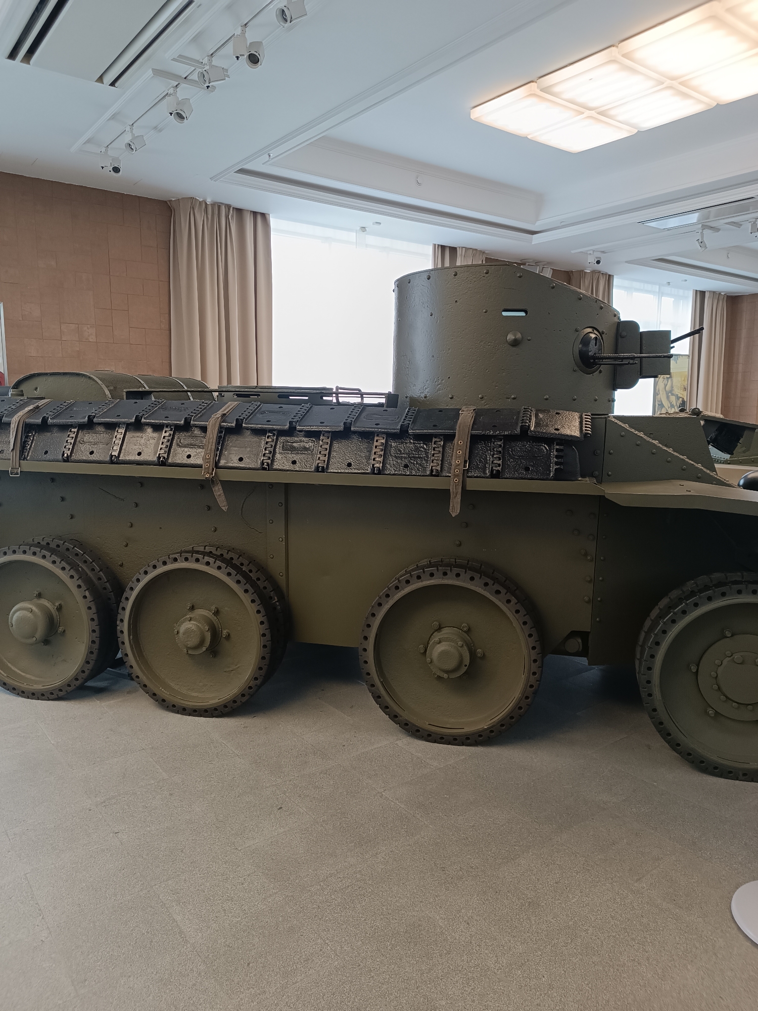 An olive-drab, six-wheeled armored vehicle is displayed indoors on a light-colored tiled floor, likely in a museum. The vehicle has a turret with a machine gun. The walls behind the vehicle are a textured brown, and beige curtains hang in the background. The ceiling features recessed lighting fixtures. This appears to be the Soviet BA-20 armored car, on display at the Museum of Military Technology UMMC in Verkhnyaya Pyshma, Russia.