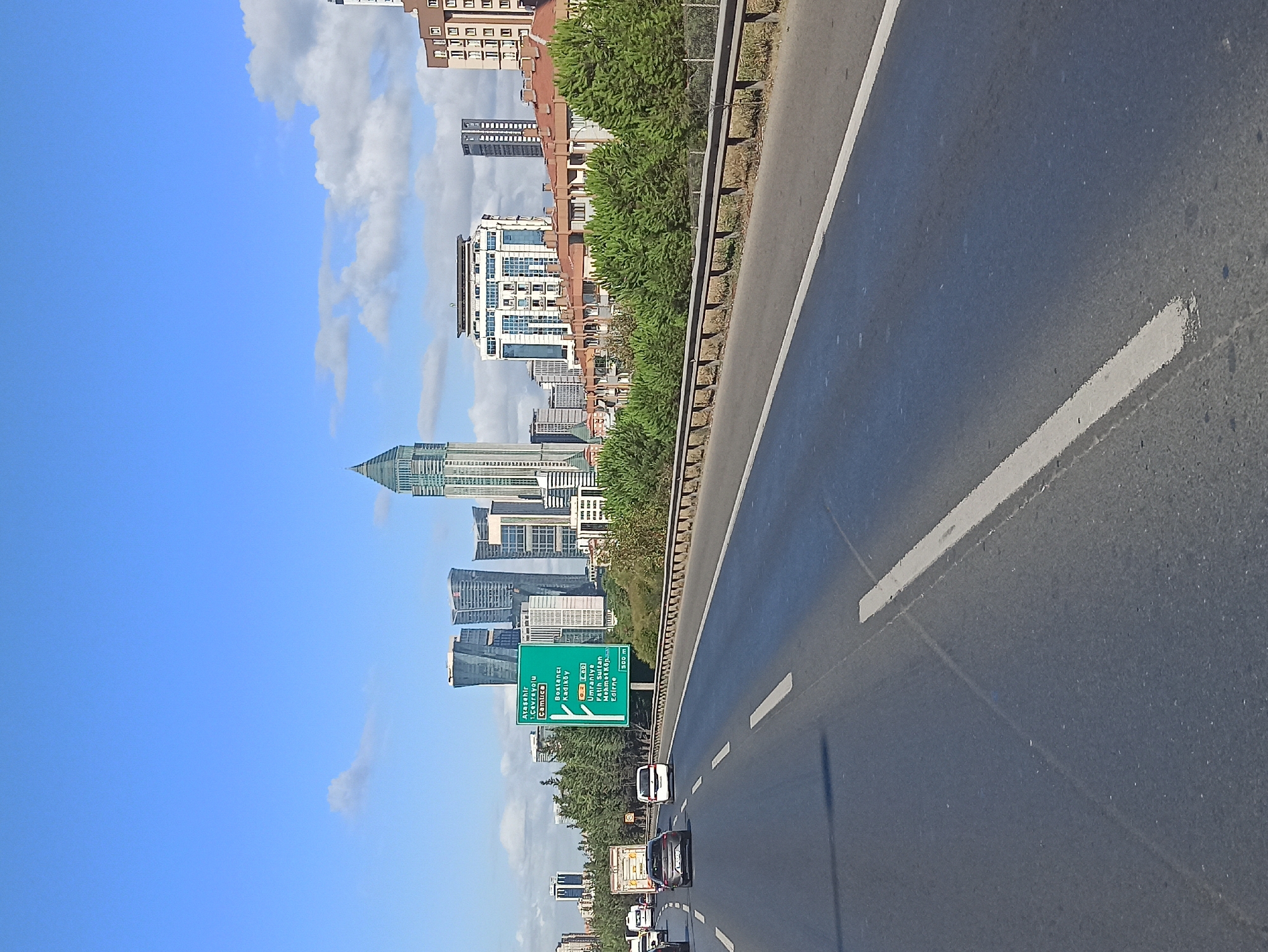 A vertical, eye-level, medium shot captures a highway in Ataşehir, Turkey, with the city's modern skyline in the background under a bright blue sky dotted with white clouds. On the right side of the road, a bright green highway sign displays directions to various locations, including "Ataşehir," "Çamlıca," "Bağdat Caddesi," "Bostancı," "Kadıköy," "Ümraniye," "Fatih Sultan Mehmet Köprüsü," and "Edirne." Several cars are visible on the highway, with a silver sedan in the foreground on the left lane and a white minivan in the right lane. A large truck is further down the highway. The road is asphalt with white lane markings. In the background, a line of trees separates the highway from a cluster of buildings, which includes low-rise residential structures with red-tiled roofs and several modern skyscrapers, some with unique architectural designs. The overall scene depicts a typical urban highway environment in a bustling Turkish city.