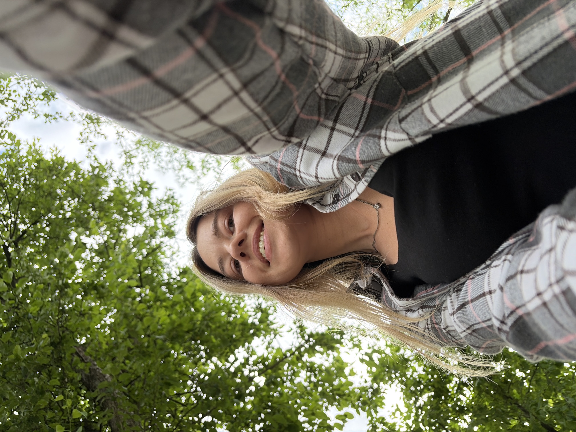 A low-angle, close-up shot captures a young woman smiling upwards, her face illuminated by the sun filtering through green leaves overhead. She is outdoors, with a blurred background of trees and sky. The woman has long, blonde hair and is wearing a black top and an open, gray and white plaid shirt with pink accents. A delicate silver necklace with a small pendant is visible around her neck. The perspective suggests she might be holding the camera or phone herself, taking a selfie. The environment is natural and appears to be a park or wooded area, which could be located in Chaikovsky, Russia.