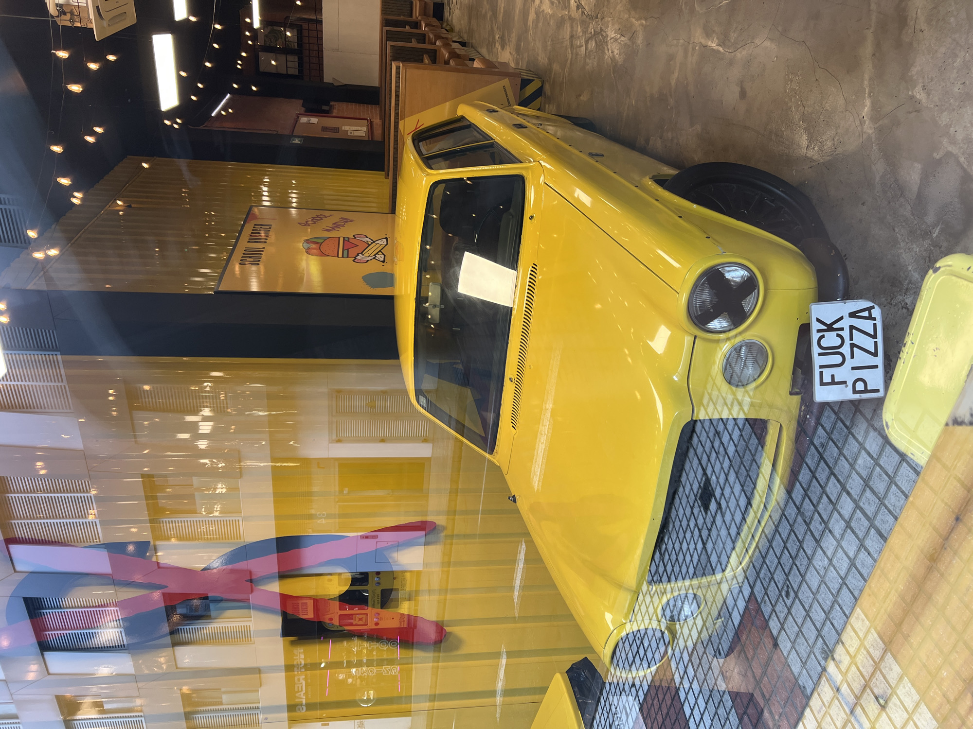 A vibrant yellow vintage car, resembling a classic Fiat 500 or similar compact model, is the central subject of this indoor scene. The car is displayed, possibly as decoration, within what appears to be a restaurant or commercial establishment in Las Palmas, Spain. The vehicle is angled towards the viewer's left, showcasing its front end, including the round headlights and grille.
A distinctive white license plate is attached to the front of the car, oriented vertically, and clearly reads "FUCK PIZZA". Behind the car, a yellow corrugated metal wall features a poster with the text "SCHOOL BURGER" prominently displayed, accompanied by a cartoon illustration of a burger wearing a graduation cap. Below "SCHOOL BURGER," the word "MANDAY" is partially visible.
The setting exhibits an industrial-chic aesthetic with numerous exposed filament light bulbs hanging from the ceiling. Reflections on a glass surface, through which the image was taken, overlay parts of the scene, particularly on the left, where a large, multi-colored abstract design (possibly another part of the decor or a reflection of external signage) is visible. The floor beneath the car is a gray and black tiled pattern. The overall impression is a quirky and themed dining space, likely a burger joint, given the prominent "SCHOOL BURGER" signage. The lighting suggests an interior setting, likely daytime, with artificial lights contributing to the ambiance.