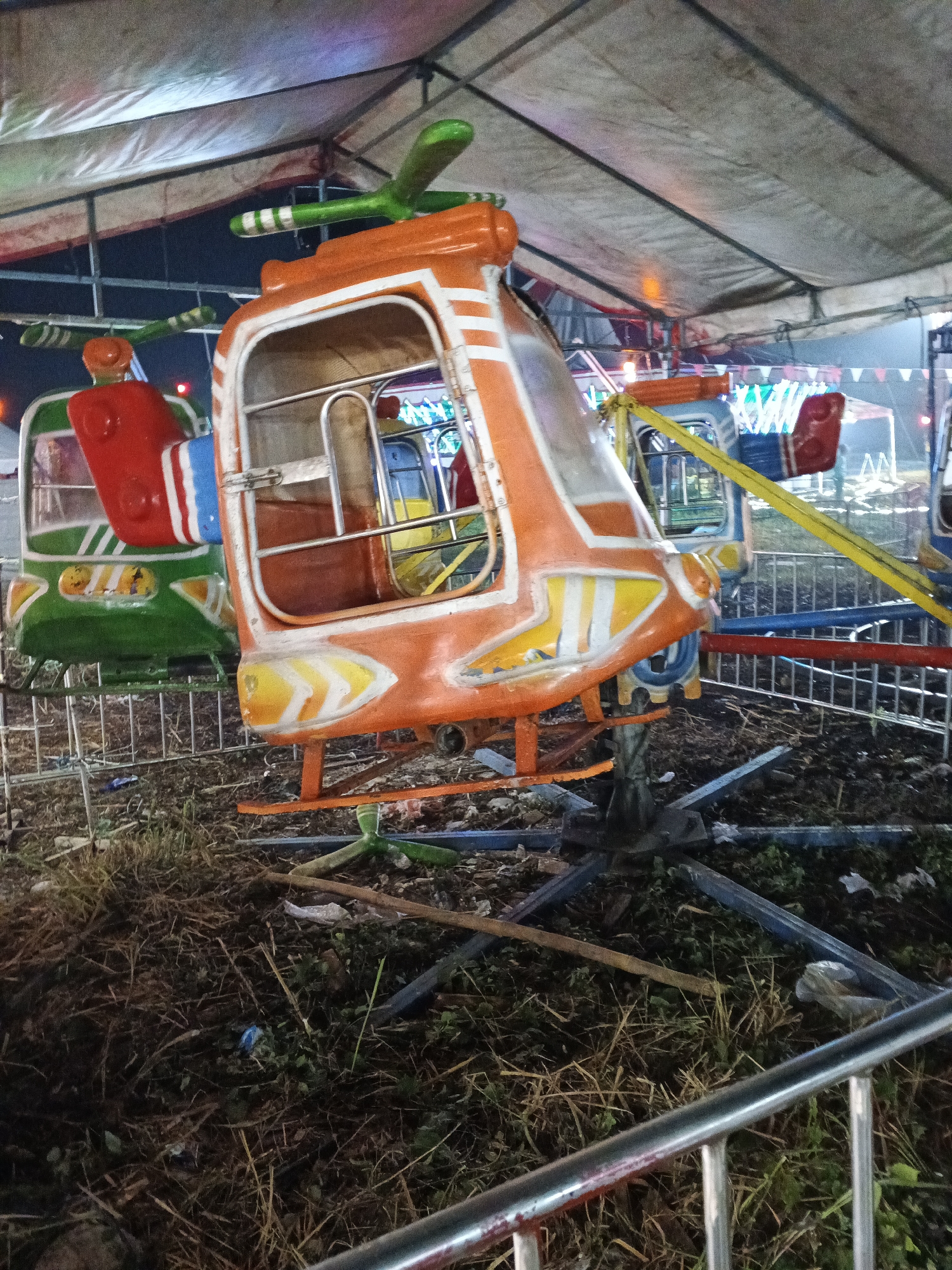 The image shows a children's amusement ride at night. The ride consists of several small helicopter-shaped vehicles attached to a central rotating mechanism. The helicopter in the foreground is orange with white stripes, and the helicopter behind it is green and red. The ride appears to be set up outdoors, possibly at a local fair or carnival, with a tented roof providing overhead cover. The ground is covered in dry grass, debris, and some scattered trash, suggesting a somewhat unkempt environment. The lighting is dim, with some artificial lights visible, indicating that it is nighttime. The overall impression is of a slightly rundown but operational amusement attraction. The location is identified as Sukabumi City, Indonesia.