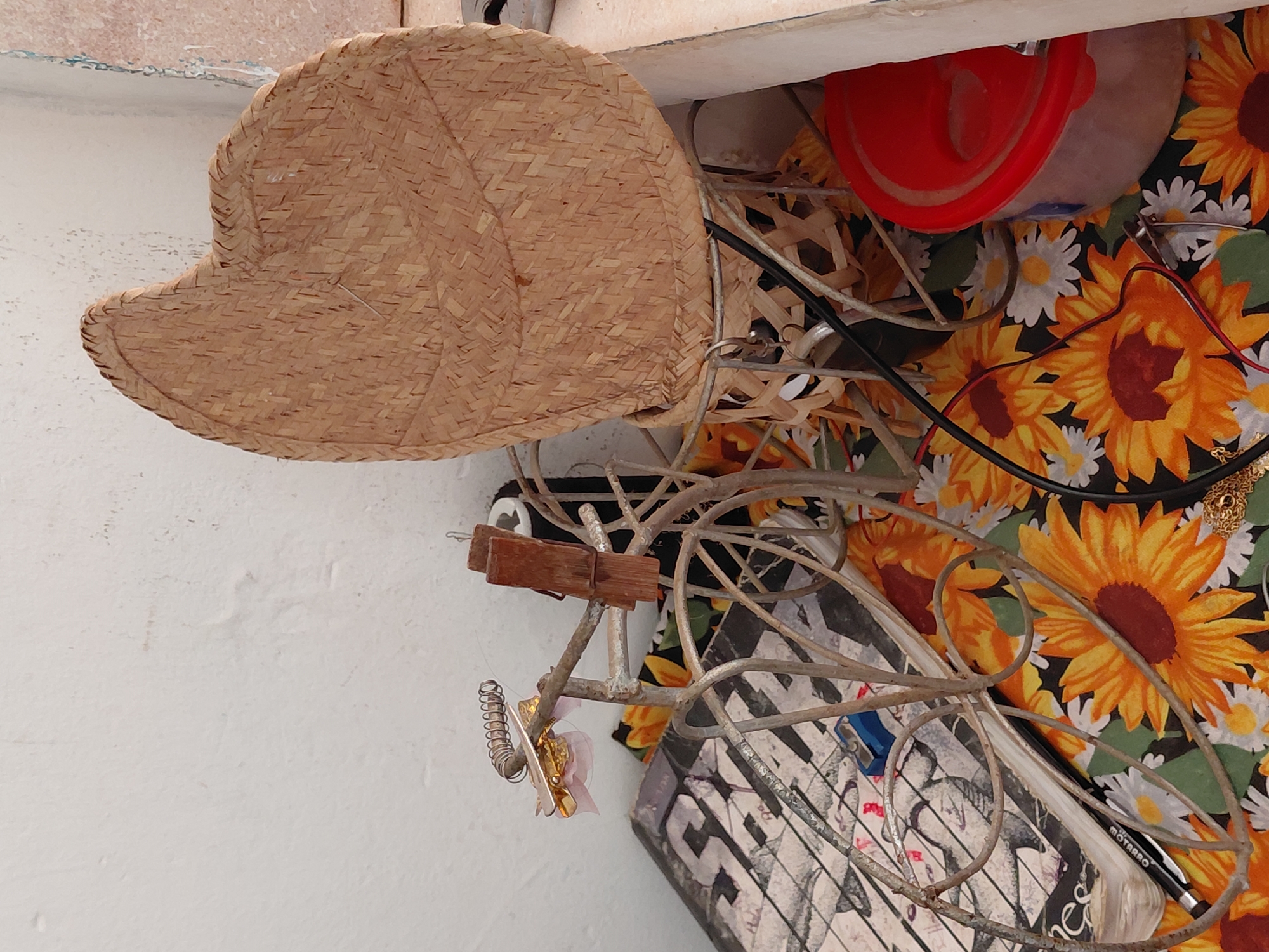 The image captures a cluttered arrangement of various objects, likely stored on a shelf or surface indoors. The scene is a static still life, with no activity or event taking place.
A large, light brown woven straw hat, displaying a prominent diagonal weave pattern and a braided edge, dominates the upper left portion of the frame. Below it and extending to the center is a decorative metal sculpture, resembling an abstract bicycle, crafted from thin, intertwined metal rods with a rustic finish. Attached to this sculpture are two small wooden clothespins and a tiny, coiled spring ornament.
Underneath and around the metal sculpture, a vibrant fabric serves as a backdrop, featuring a bold pattern of yellow and orange sunflowers interspersed with white daisies on a dark background. Nestled among these items is a translucent plastic container with a bright red lid, partially visible. Black electrical cables or wires are also interwoven among the objects, suggesting proximity to electronic devices.
On the right side of the image, resting on the sunflower fabric, is a thin gold-colored chain or piece of jewelry. Further down, a black pencil or eyeliner with the word "MOTARRO" clearly visible on its side lies alongside a small blue plastic pencil sharpener. Parts of a book cover or a piece of paper with black and white text, including the letters "SA," are discernible in the lower center.
The lighting is soft and diffuse, indicating an indoor setting or a heavily shaded area, with no strong shadows to suggest a specific time of day or outdoor weather. The collection of items suggests personal storage or an accumulation of everyday objects. While the location is Santa Ana, Venezuela, there are no specific visual cues within the image to explicitly confirm this.