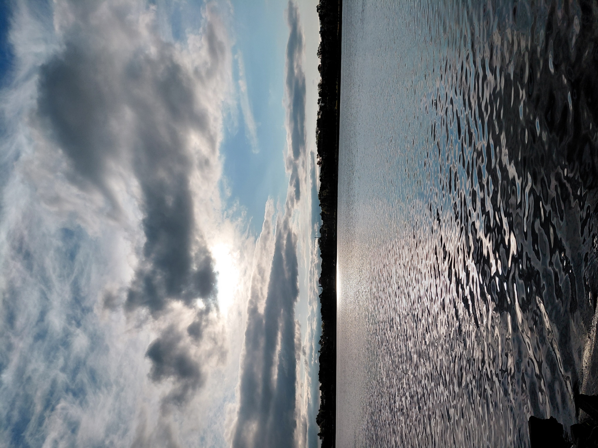 This is a landscape photograph taken at dusk or dawn, overlooking the Paraná River in Encarnación, Paraguay. The foreground features the dark, rippling surface of the river, with some dark rocks visible on the left edge of the frame. The middle ground is dominated by a silhouetted treeline along the opposite bank, with some distant structures and lights indicating a populated area. The sky above is filled with dramatic clouds, a mix of dark, heavy formations and lighter, wispy ones, with patches of blue sky visible. The sun is partially obscured by clouds, casting a bright, reflective sheen on the water's surface. There are no people or animals visible in the image.