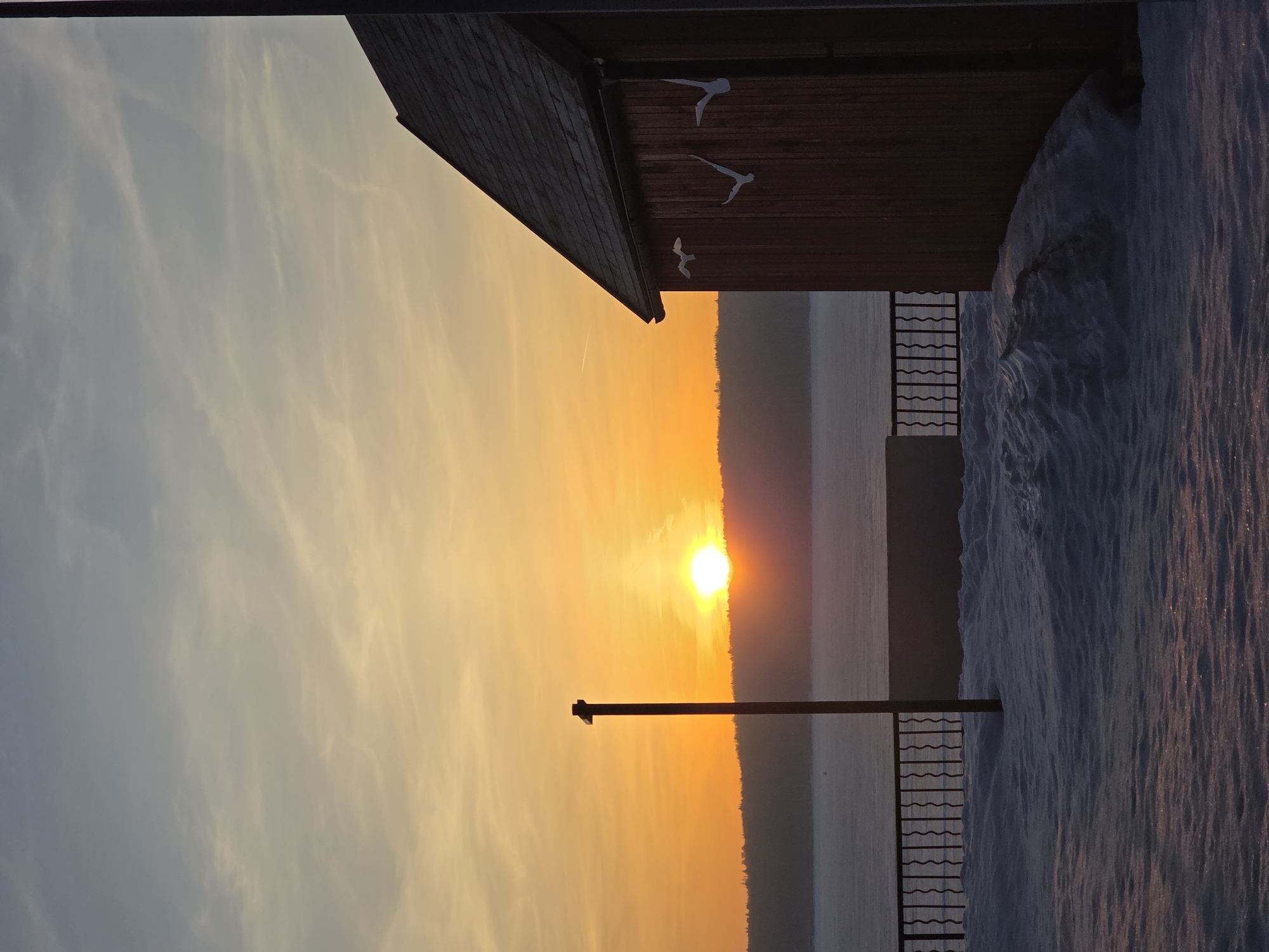The image shows a sunset over what appears to be the Kama River in Chaikovsky, Russia, evidenced by the presence of typical Russian architectural features like wooden structures and metal railings seen on the riverbank. The sun is setting on the horizon, casting a warm orange glow across the sky and water. Snow covers the ground, indicating a winter scene. A wooden structure with decorative bird cutouts is partially visible on the upper right side of the frame. The sky transitions from a soft blue to a vibrant orange as the sun nears the horizon. A lamppost is silhouetted against the bright sunset.