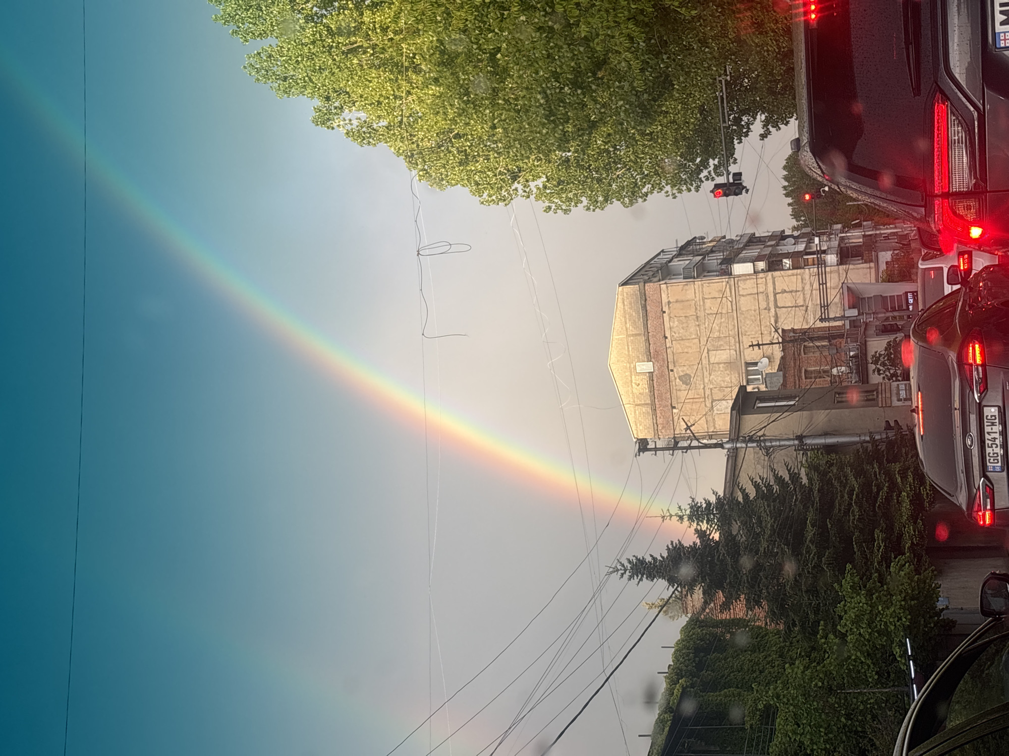 The image captures a partial rainbow arcing across the sky in what appears to be an urban environment. The scene is shot from a low angle, likely from within a vehicle, evidenced by the car’s interior visible on the bottom right. In the foreground, several vehicles are stationary, indicated by the illuminated brake lights. Car’s plate is GG-541-WG. A traffic light overhead shows red. Buildings of varying heights and architectural styles are visible, along with dense trees. Overhead power lines cut across the sky. Based on the license plate seen, the photo was likely taken in Tbilisi, Georgia.